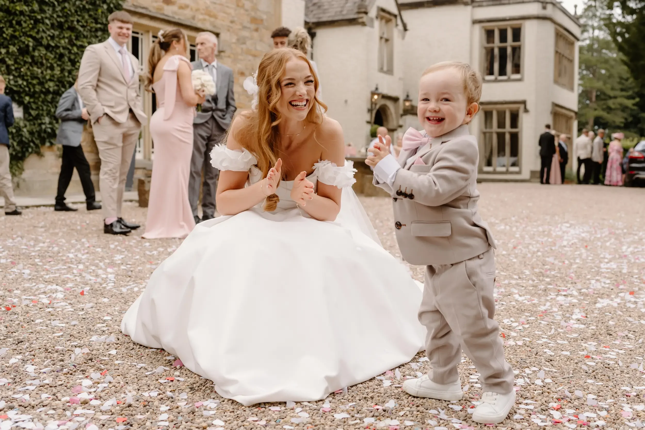 A woman in a wedding dress kneeling on the ground, joyfully interacting with a young boy in a beige suit and pink bow tie, at a wedding celebration outside a stone building, with other guests in formal attire in the background.