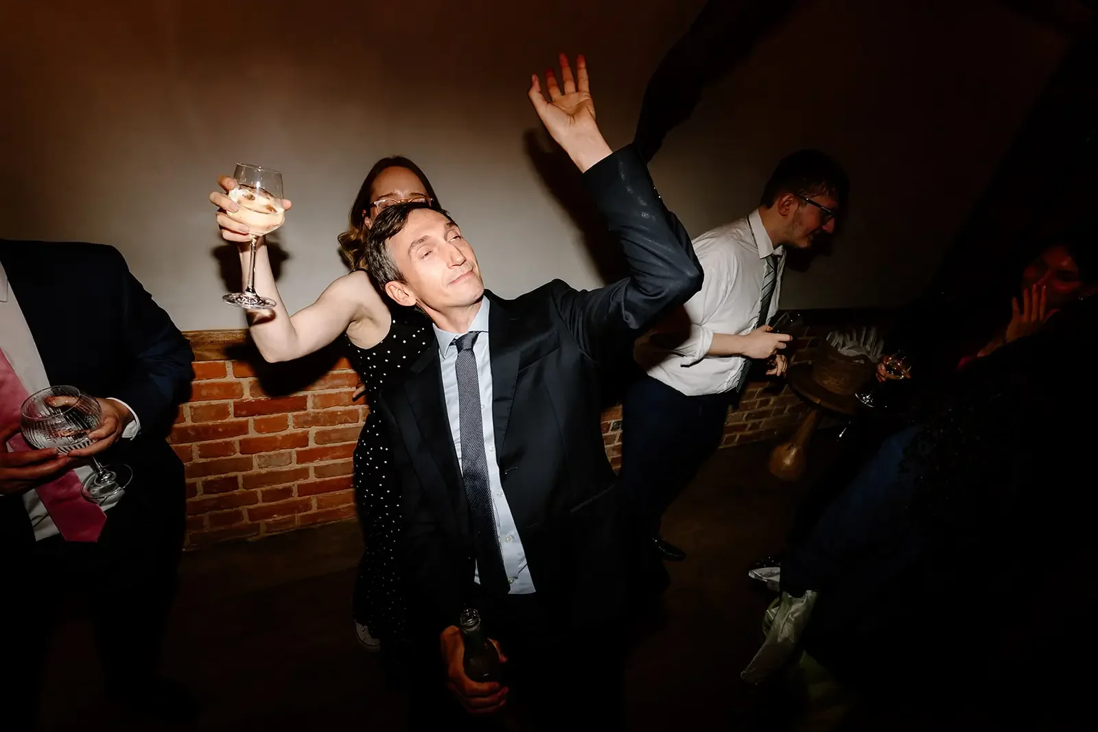 People dancing and having fun at a party, with a man in the center raising his hand and holding a drink, others holding glasses and bottles, in a room with brick walls.