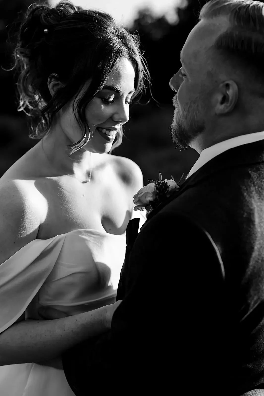 Black and white photo of a bride and groom during their wedding, close-up facing each other, smiling, with the bride wearing an off-shoulder dress and the groom in a tuxedo with a boutonnière.