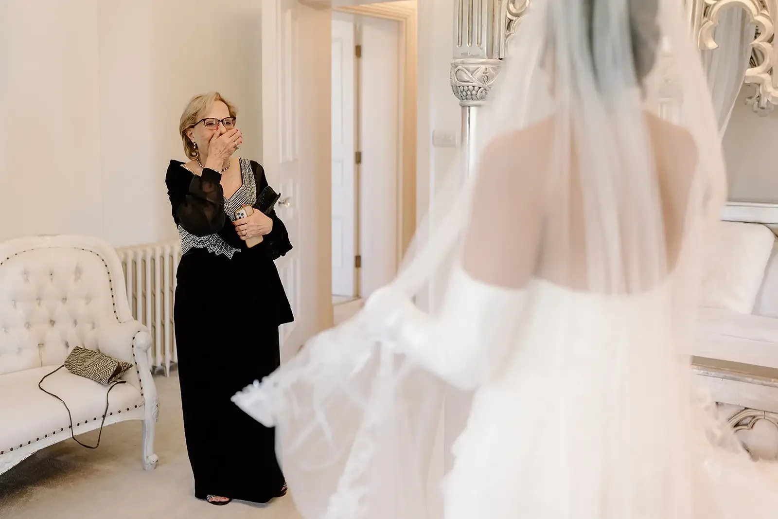 An older woman in a black dress with sheer black sleeves and glasses is standing in a room with beige walls, covering her mouth with her hand and holding something in her other hand. She appears emotional, possibly crying, while looking at someone in