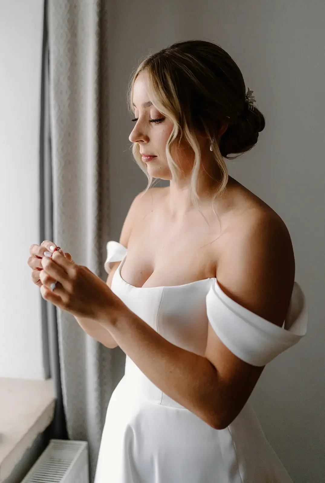 A woman in a wedding dress holding her hands together near her face, looking down, standing by a window.