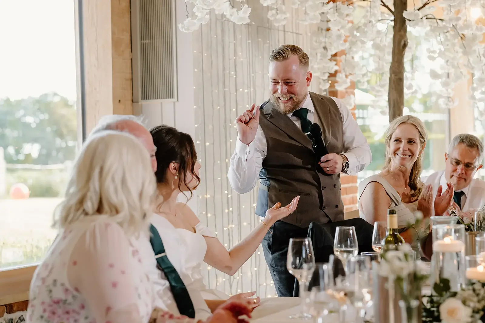 A man is giving a toast and making a speech at a wedding reception, standing with a microphone in hand and smiling, while the bridal party listens and claps.