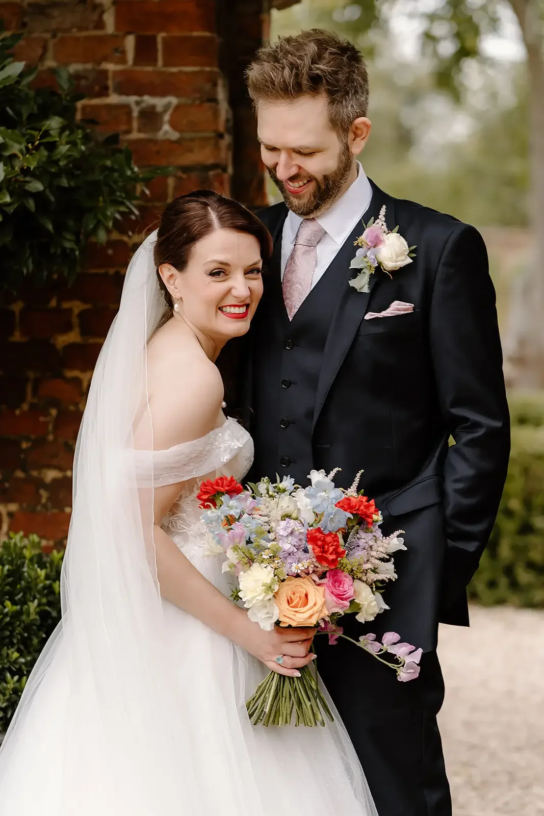 Bride and groom standing outdoors near a brick wall, smiling, with the bride holding a colorful bouquet of flowers.