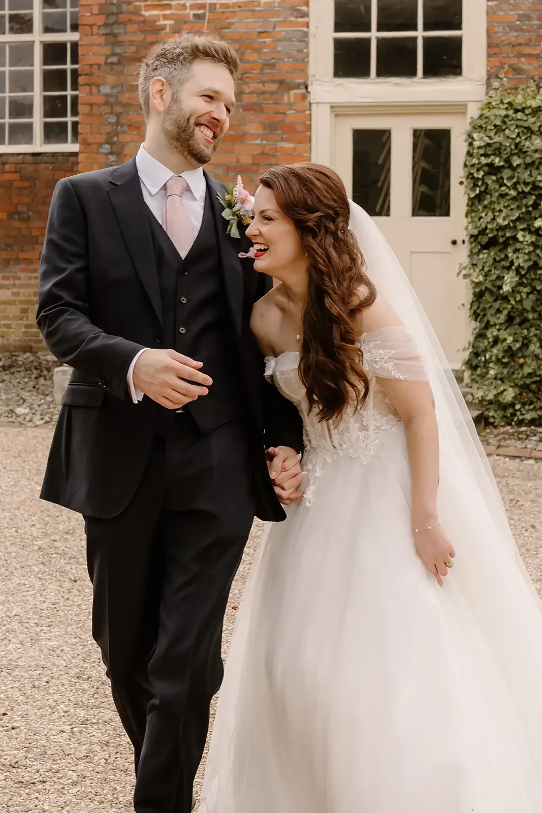 A bride and groom laughing and holding hands outside in front of a brick building.