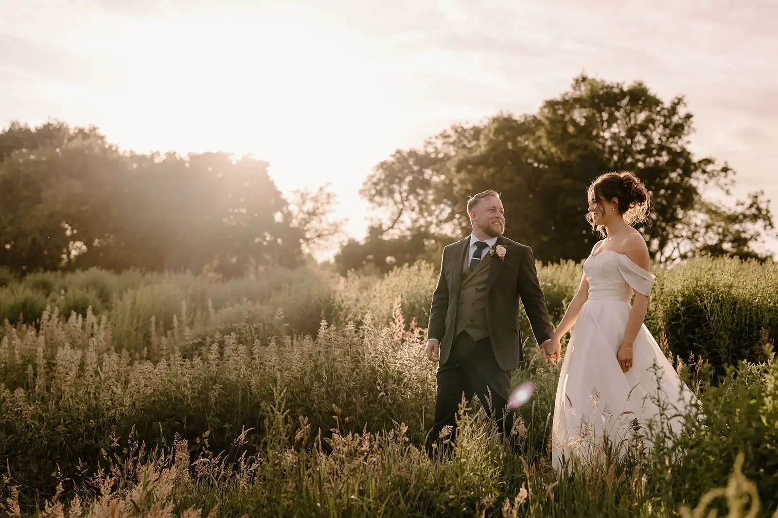 A bride and groom holding hands and smiling at each other in a field of tall grass and flowers during sunset.