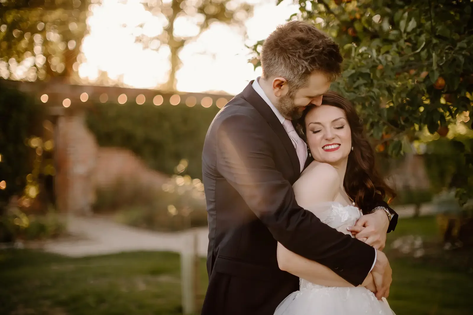 A man and woman in wedding attire embrace outdoors during sunset, smiling. The man is wearing a black suit and the woman a white wedding dress, with greenery and string lights in the background.