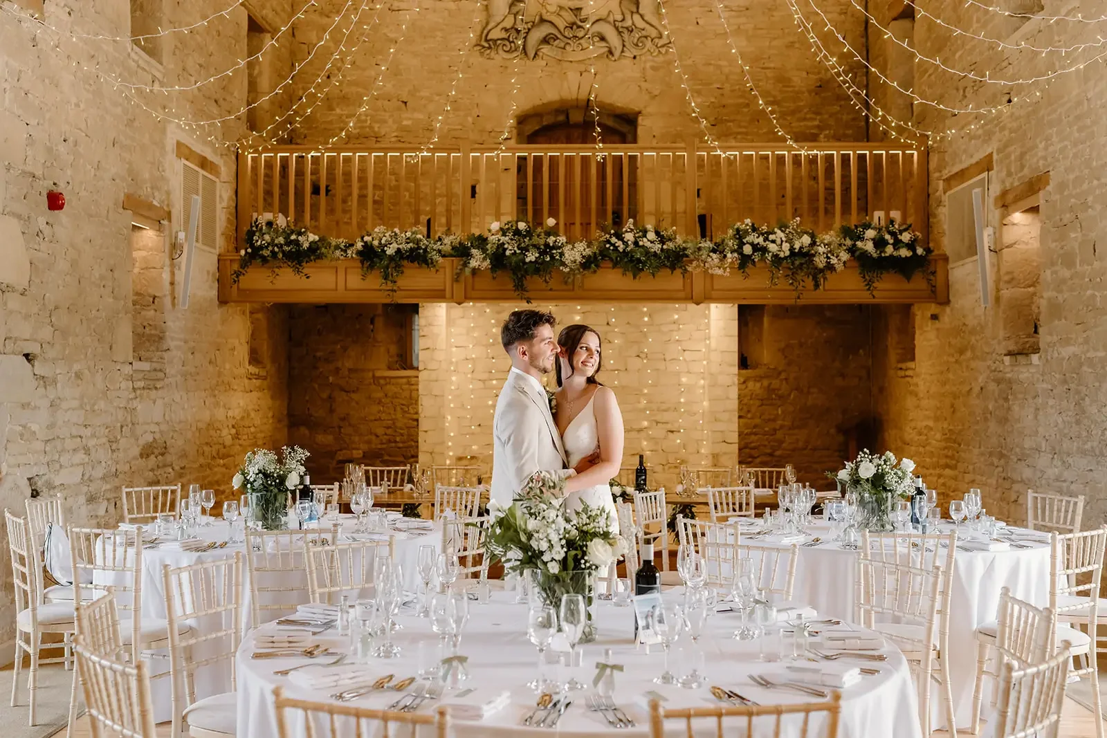 A bride and groom standing together at a wedding reception, smiling and embracing in a rustic venue with exposed brick walls, decorated with string lights and floral arrangements.