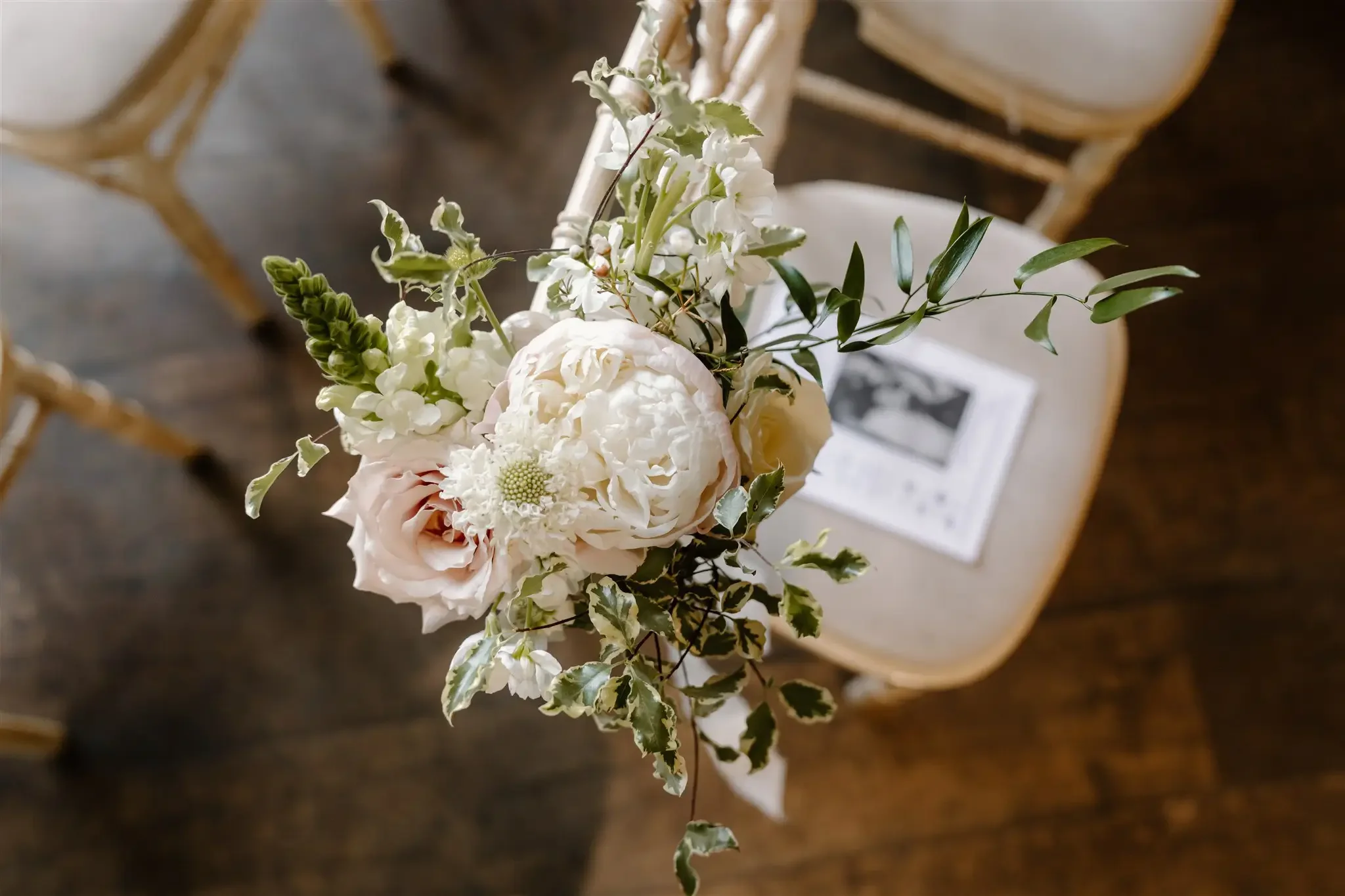 A bouquet of white and light pink flowers with green foliage on a chair with a paper on it, set on a wooden floor.
