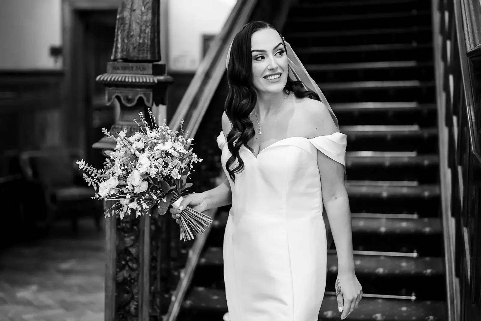 Black and white photo of a woman in an off-the-shoulder wedding dress holding a bouquet of flowers, standing on a staircase indoors.