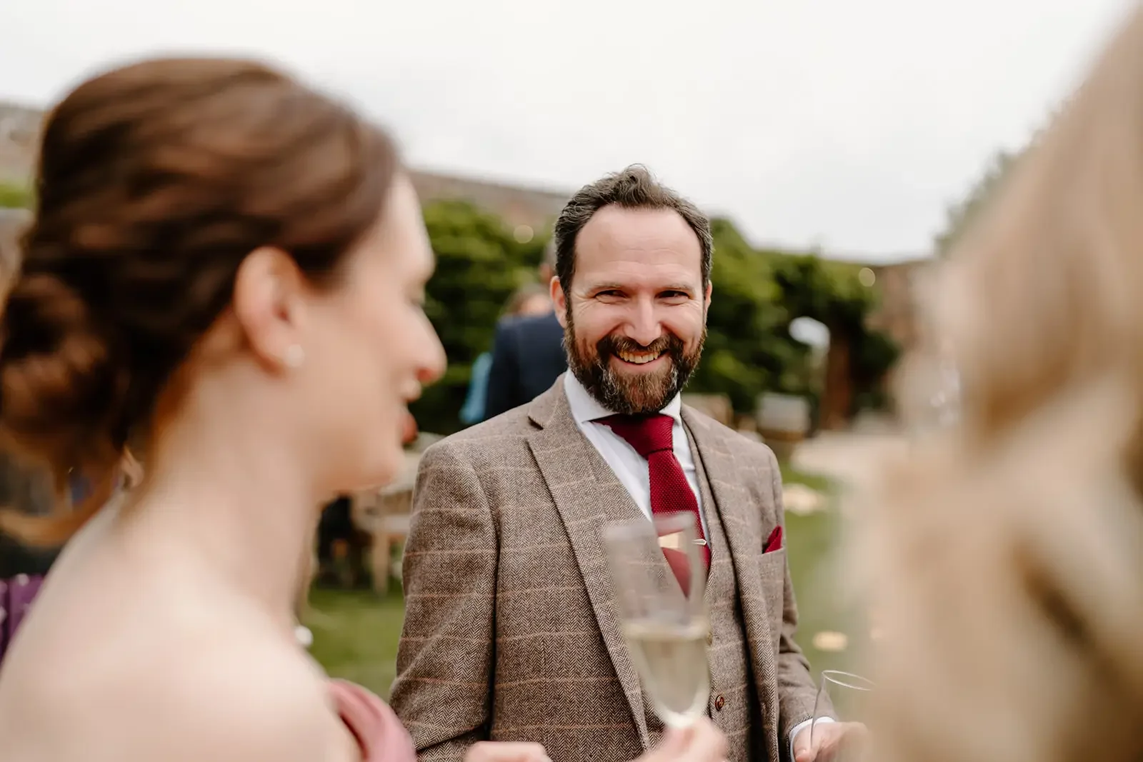 A man with a beard and a woman with brown hair in an outdoor gathering, smiling and holding champagne glasses.