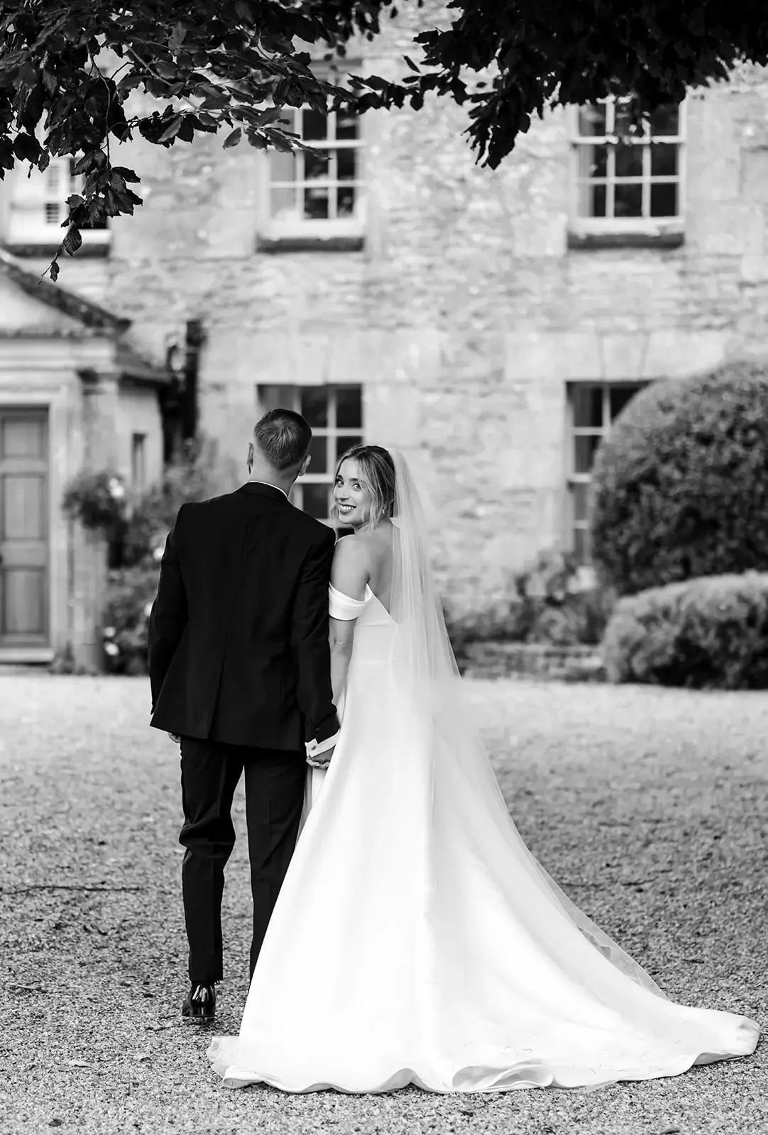A bride and groom walking away outdoors, the bride smiling and looking back, in front of a brick building, black and white photo.
