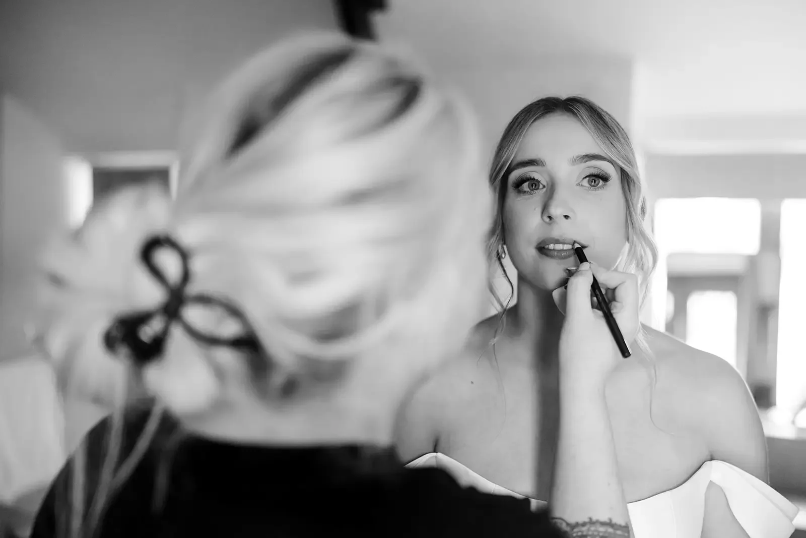 Black and white photograph of a woman getting her makeup done, with a makeup artist in the foreground but out of focus.