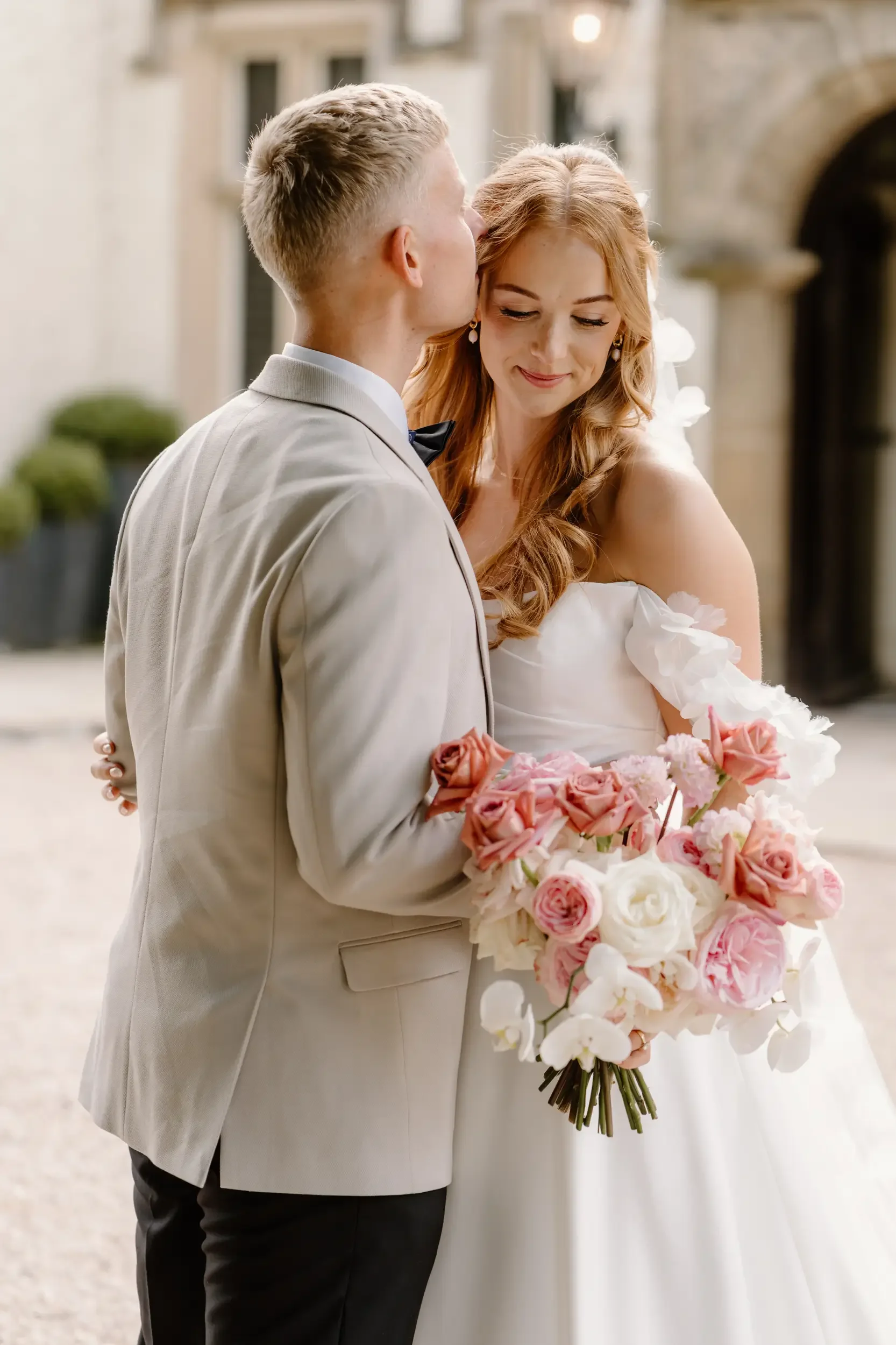 A bride and groom close together, the groom kissing the bride on the forehead, holding a bouquet of pink, white, and peach flowers.