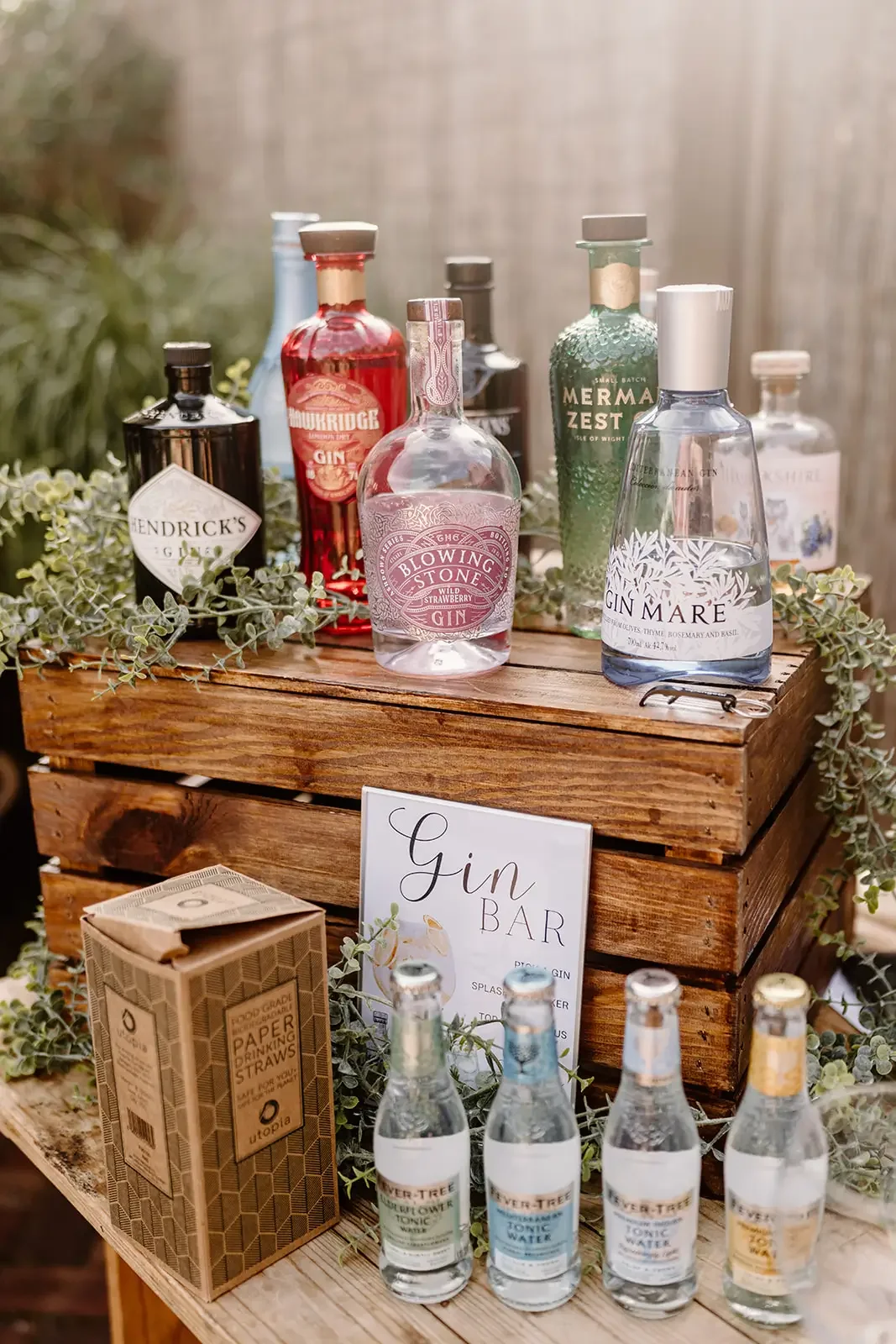 A wooden display stand with several bottles of gin, tonic water, and a carton of paper drinking straws, labeled as a gin bar, decorated with greenery and a sign.
