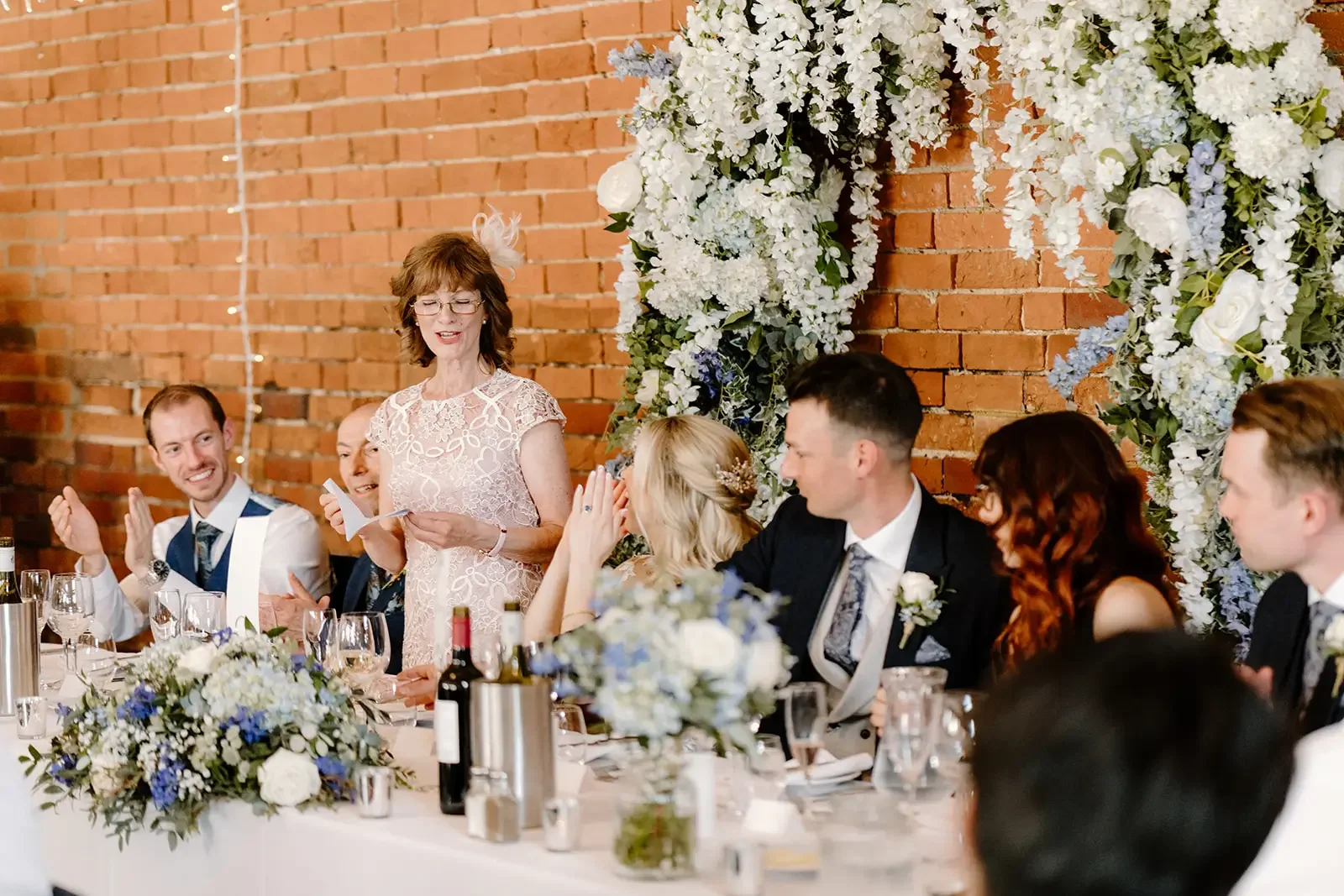 A woman giving a speech at a wedding reception with a floral backdrop and guests listening.