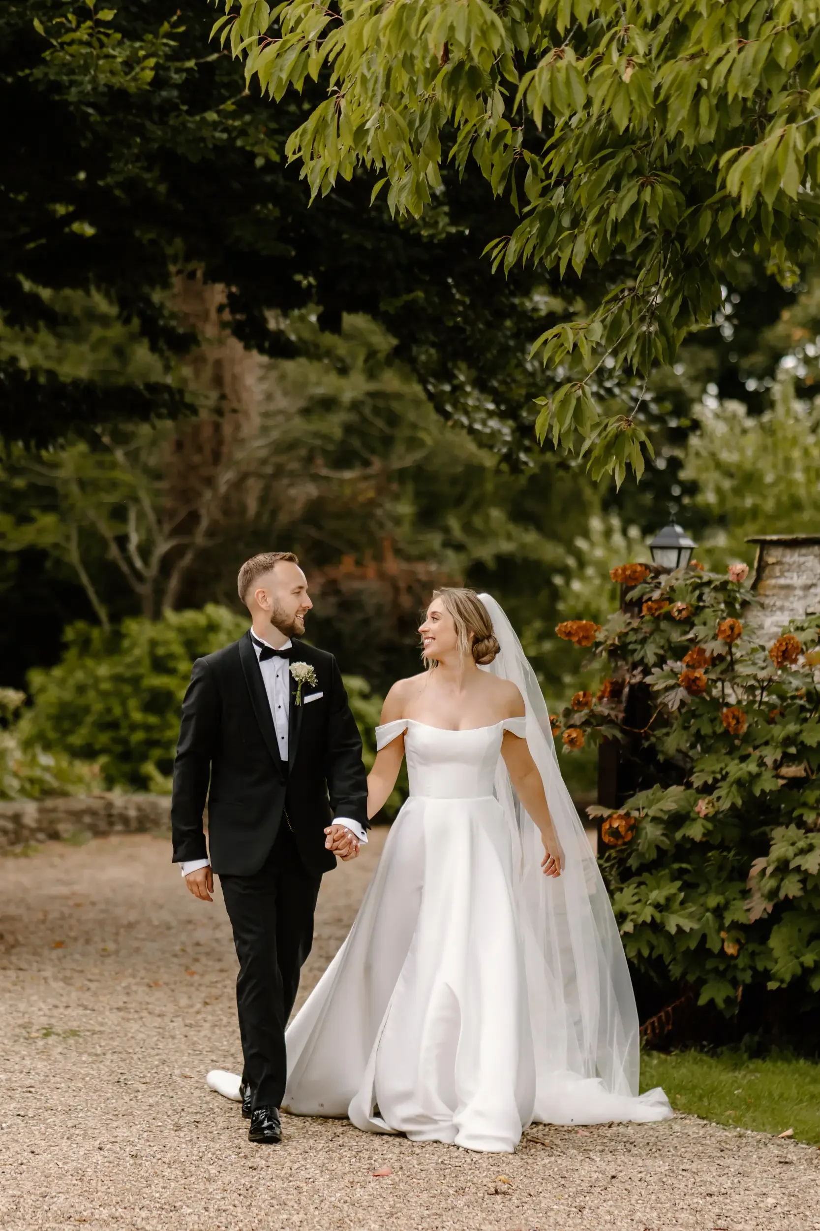 A bride and groom walking hand in hand outdoors, smiling at each other, with lush greenery and orange flowers in the background.