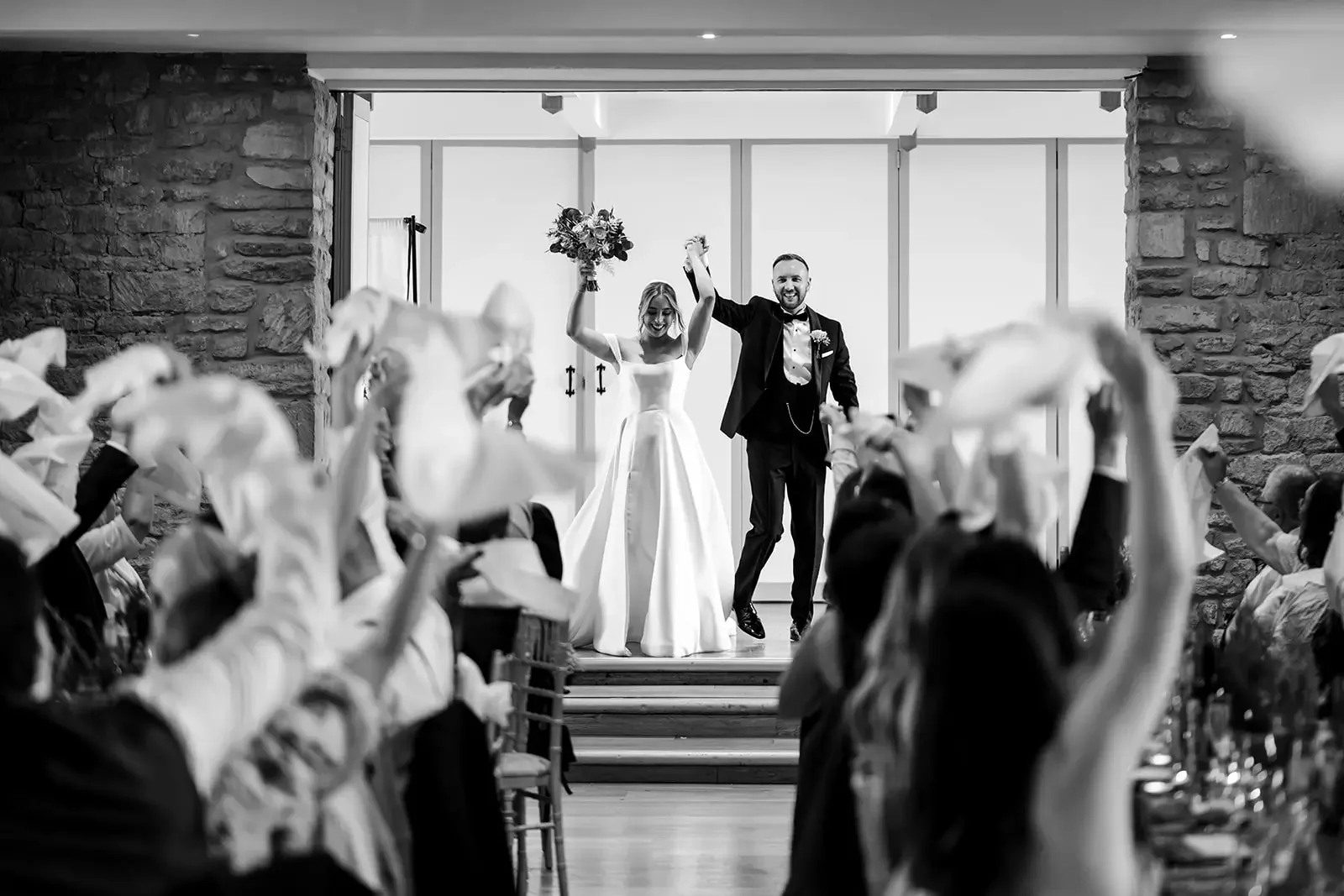 Black and white photo of a newlywed couple holding hands and smiling on a stage, with wedding guests celebrating in the foreground.