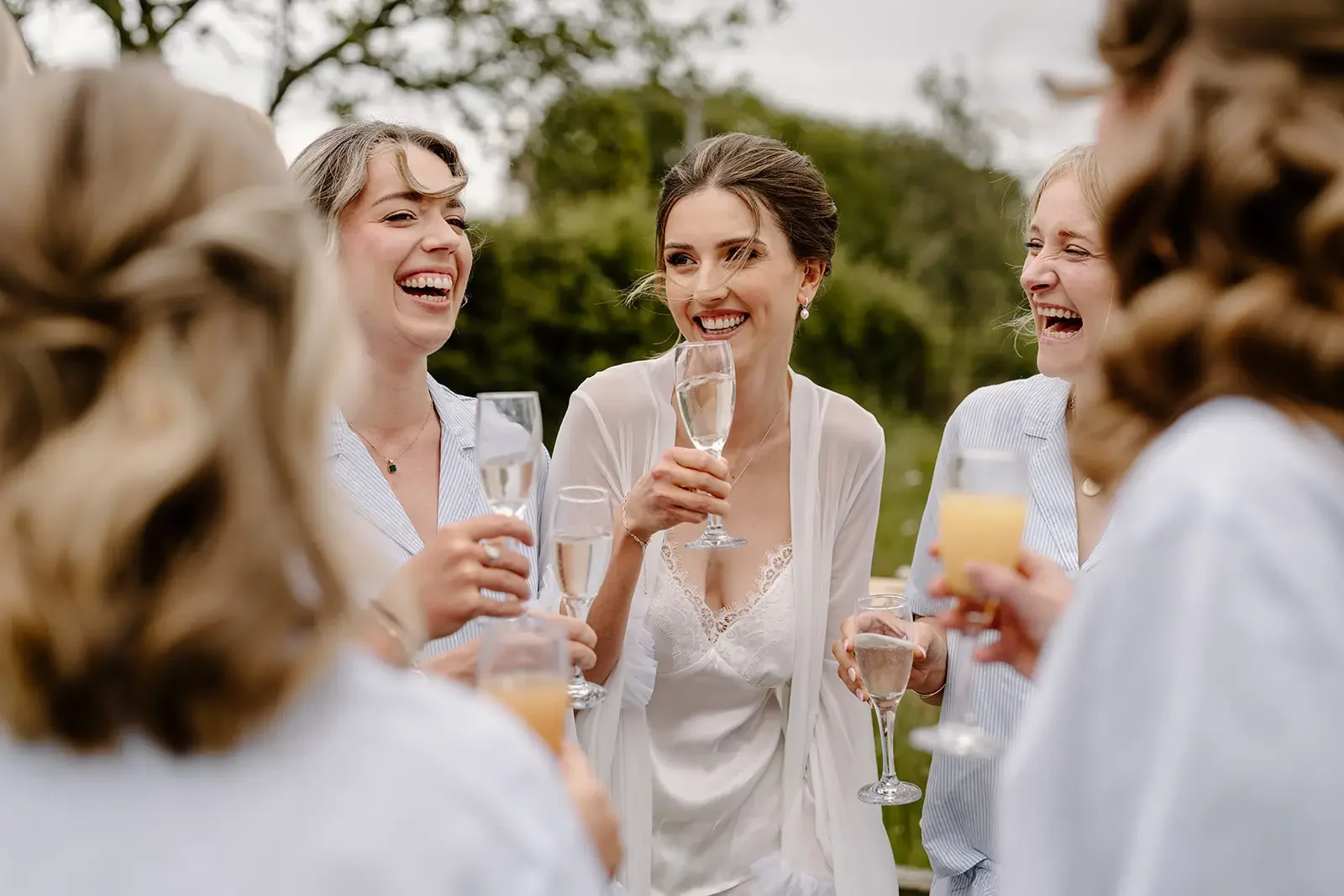 Group of women in white attire celebrating outdoors with champagne and orange drinks, laughing and smiling.