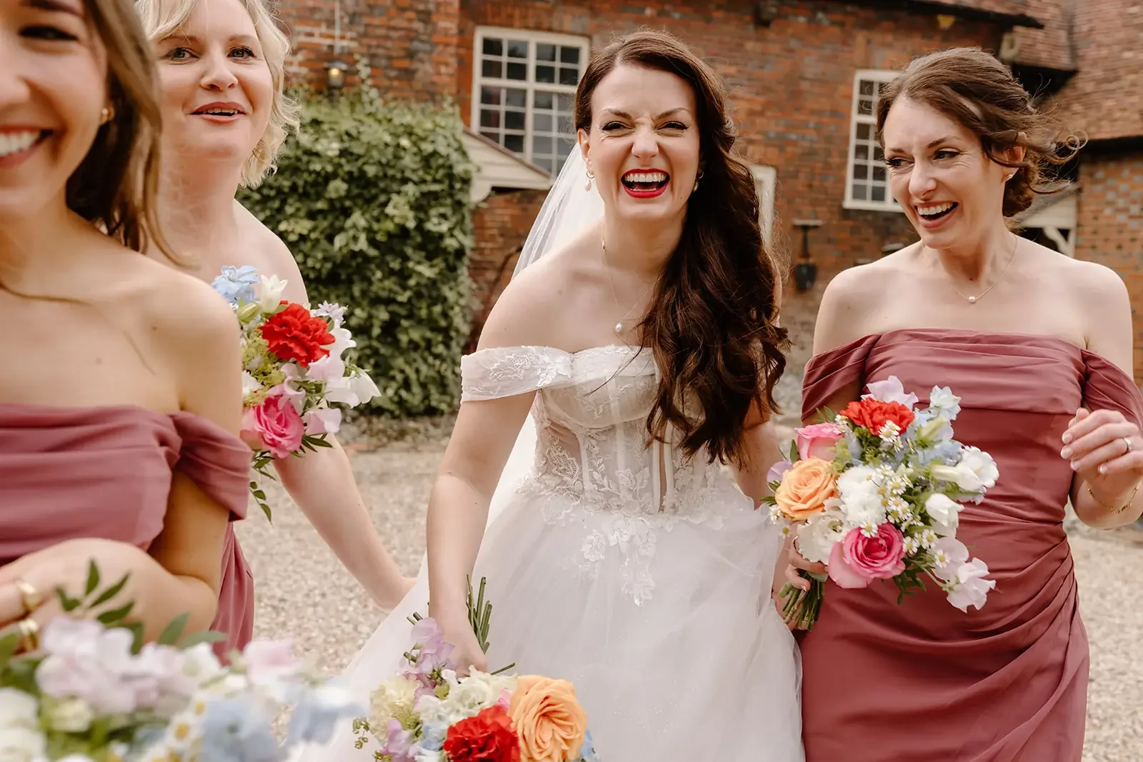 A group of women, including a bride in a white wedding dress and bridesmaids in mauve dresses, laughing and holding bouquets of flowers outside a brick building.