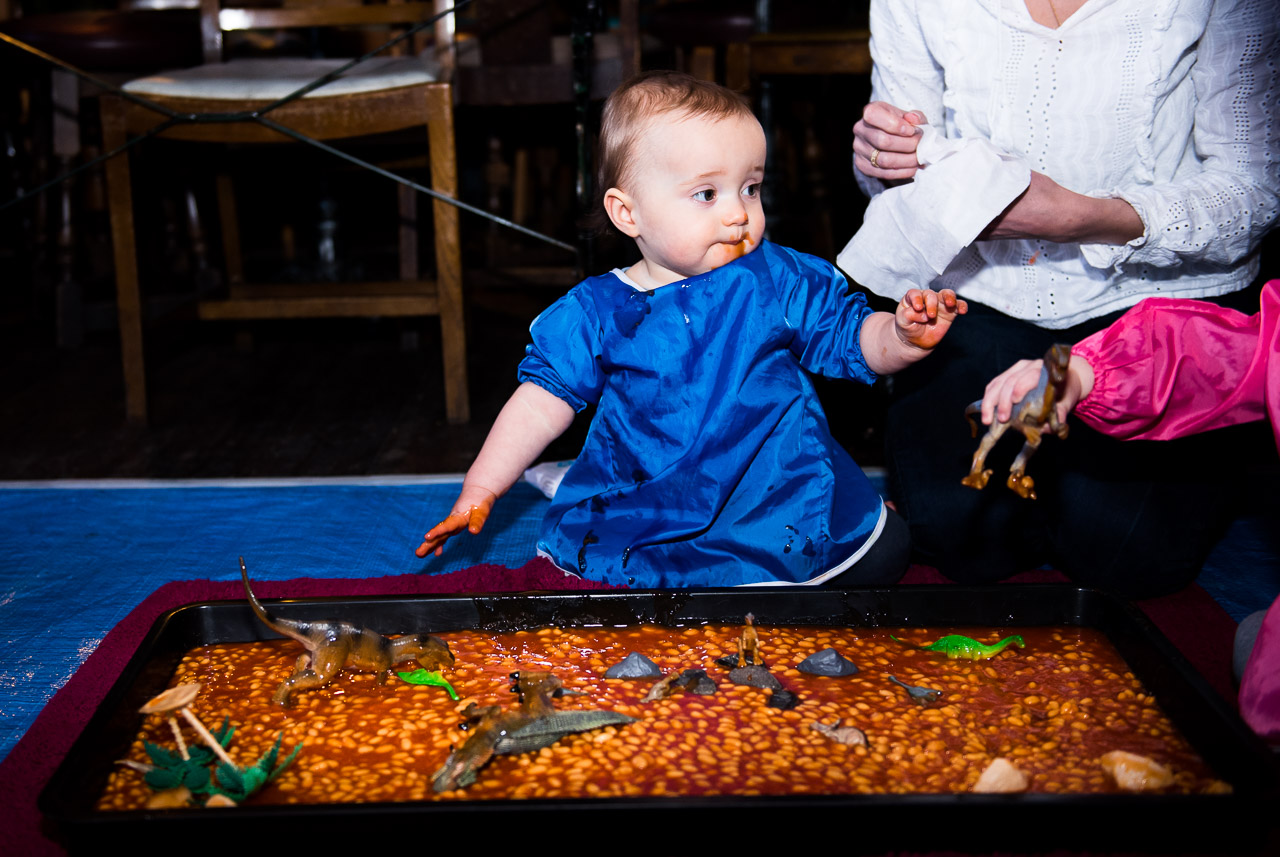  Little boy is playing in a tray that has baked beans. There are also dinosaurs in the tray to play with 