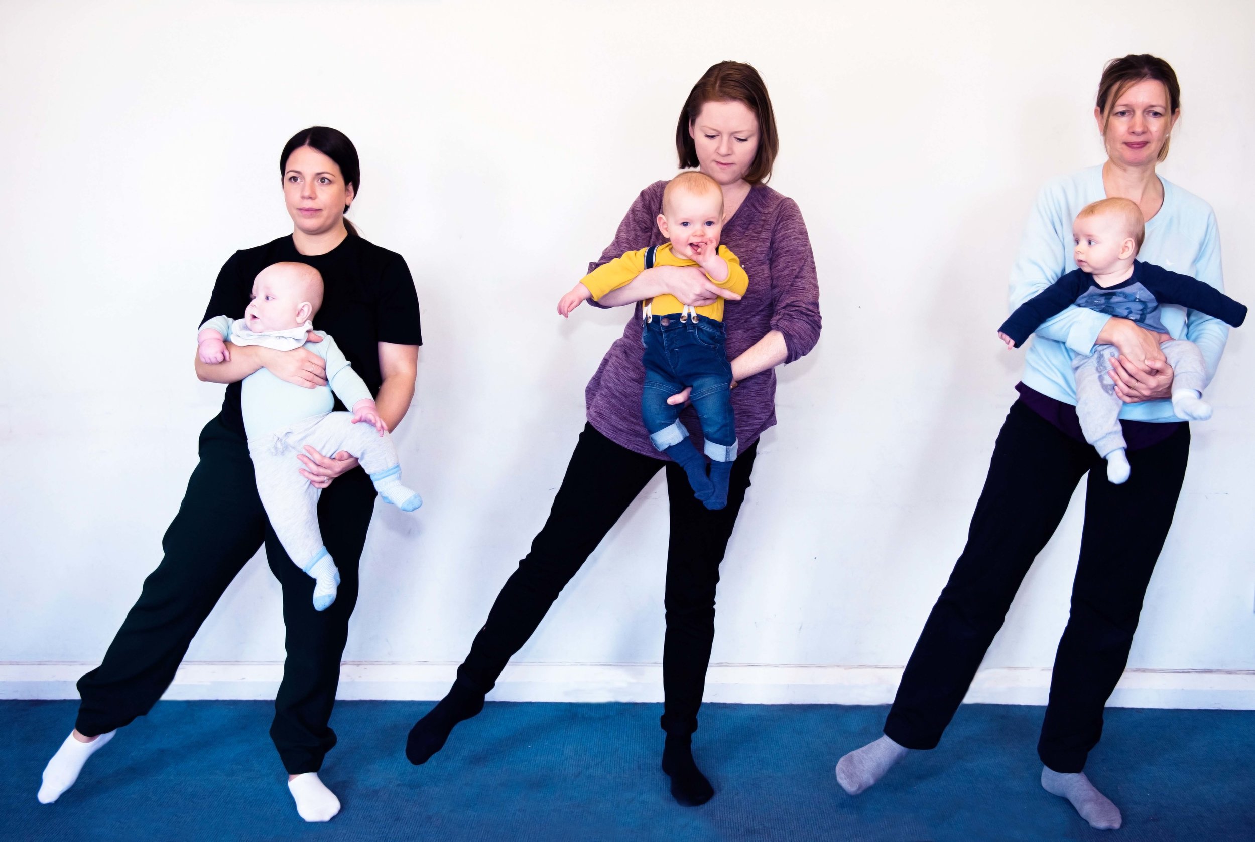  3 mums are standing in a row, holding their babies with the backs on their fronts. They have one leg straight out to the side 