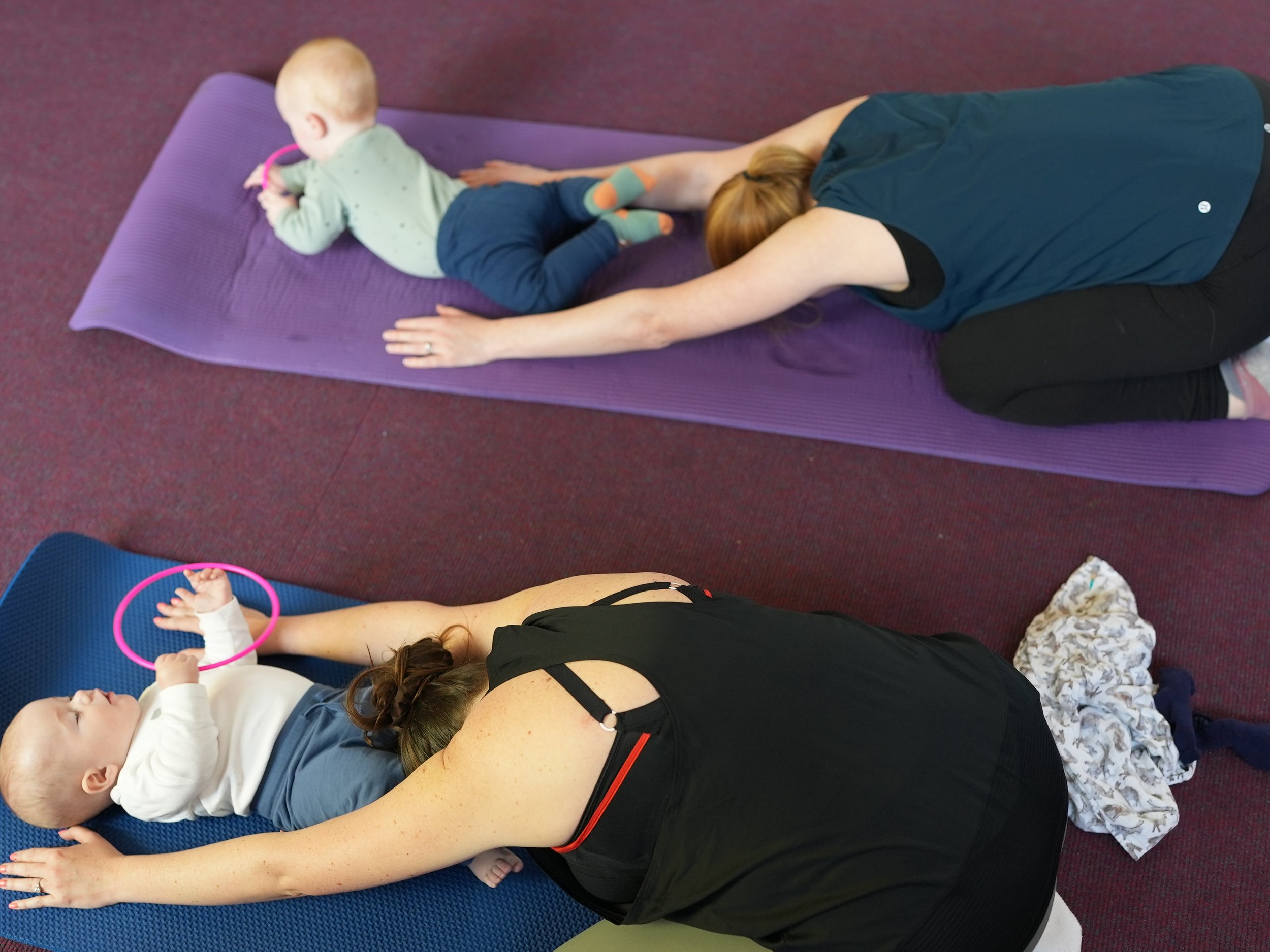  Mum is in a child’s pose with her baby near her head. The babies are being distracted with a sensory toy 
