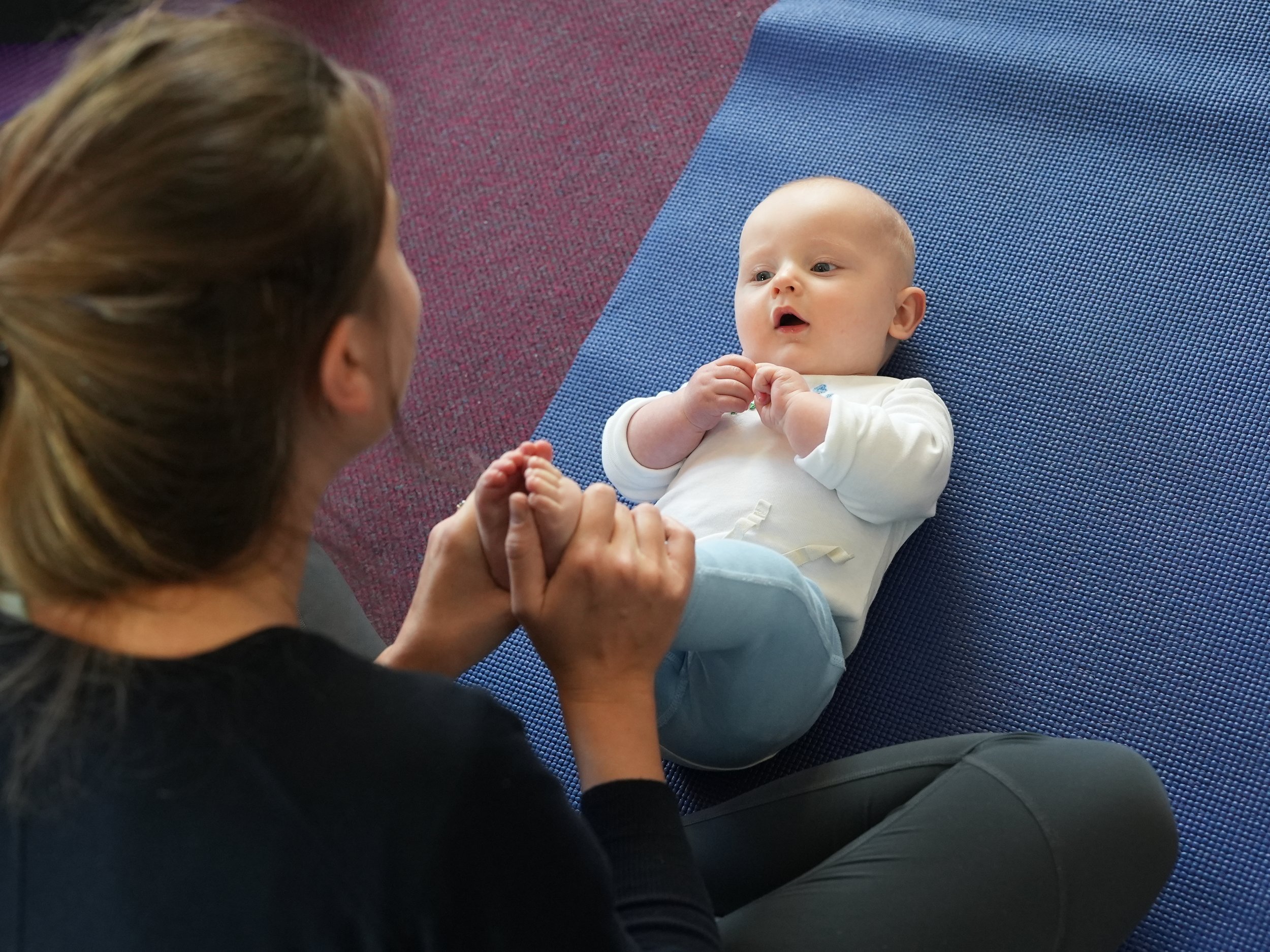 Baby boy is laying on a blue yoga mat and is looking up at his mum whilst she holds his feet