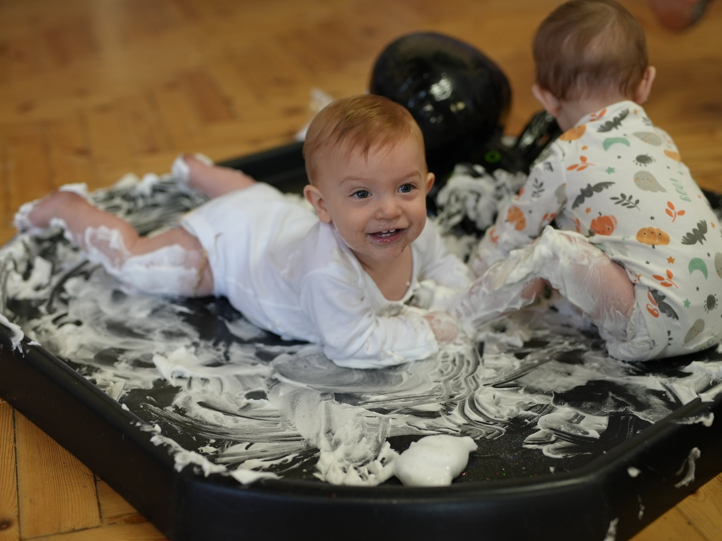  little boy is smiling whilst playing in a big tray filled with shaving foam 