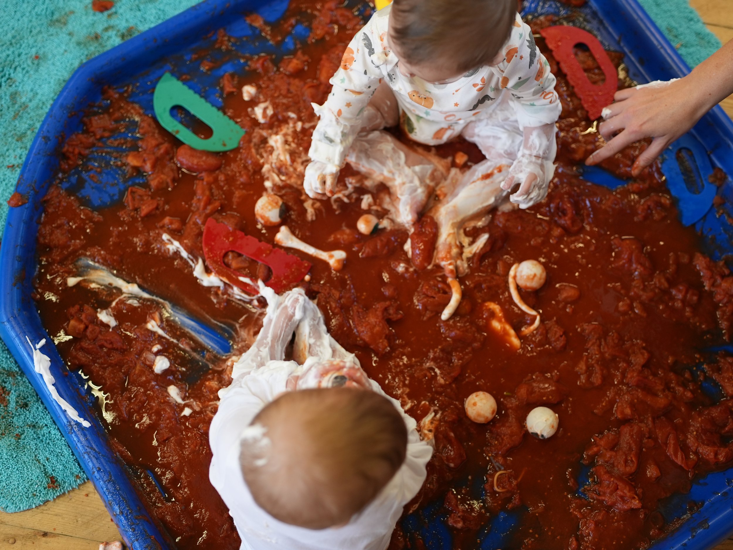 2 babies are playing in a huge tuff tray filled with tinned tomatoes