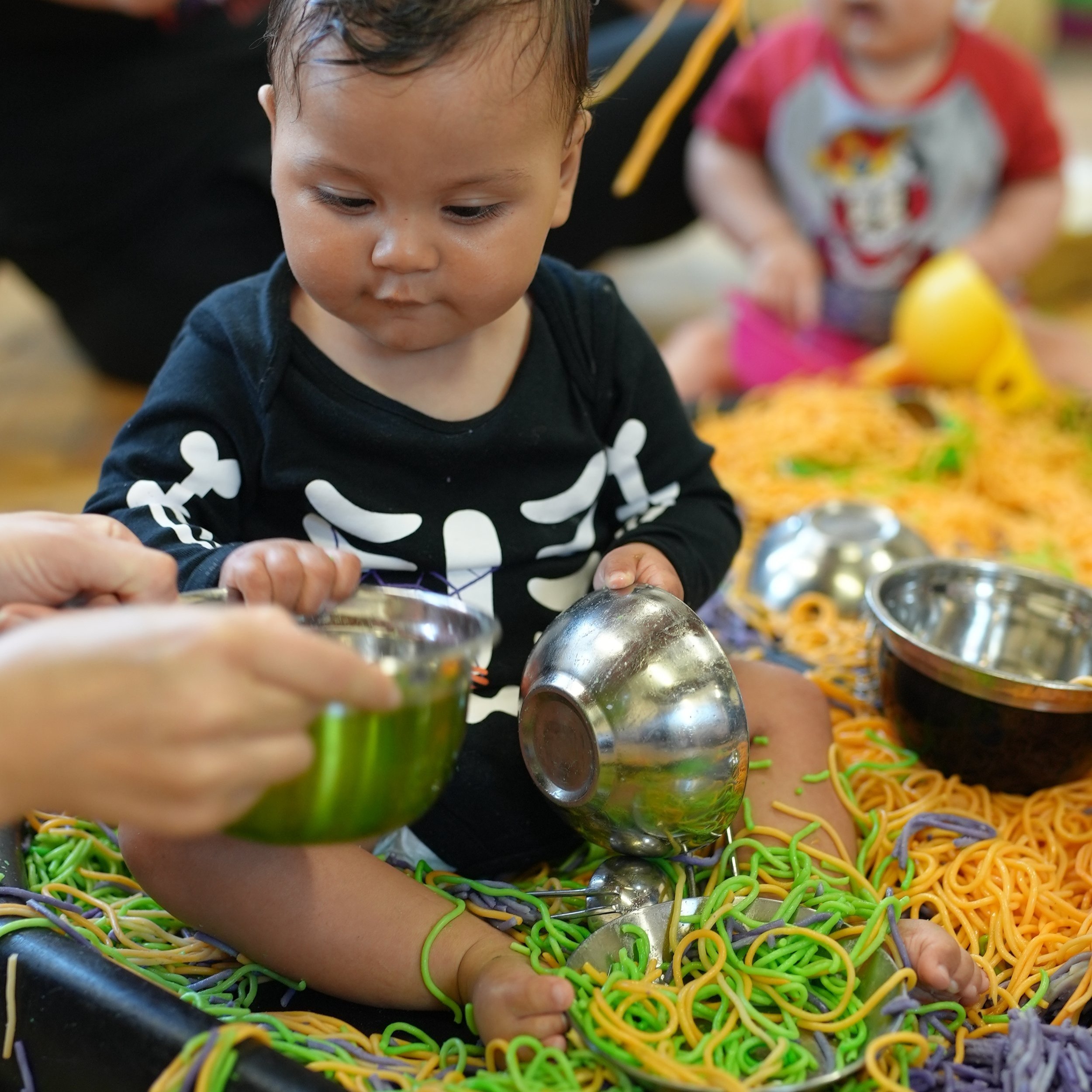  Little boy is in a huge tray filled with colourful cooked spaghetti. He is holding a metal bowl 