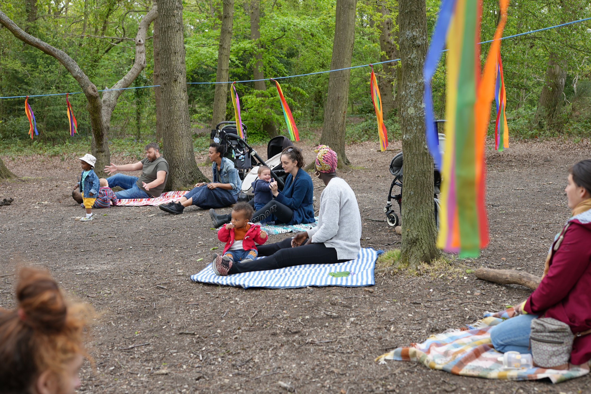  Parents have brought their own mats to the woods and are all sitting around in a bug circle. You can see rainbow ribbons hanging from the trees 