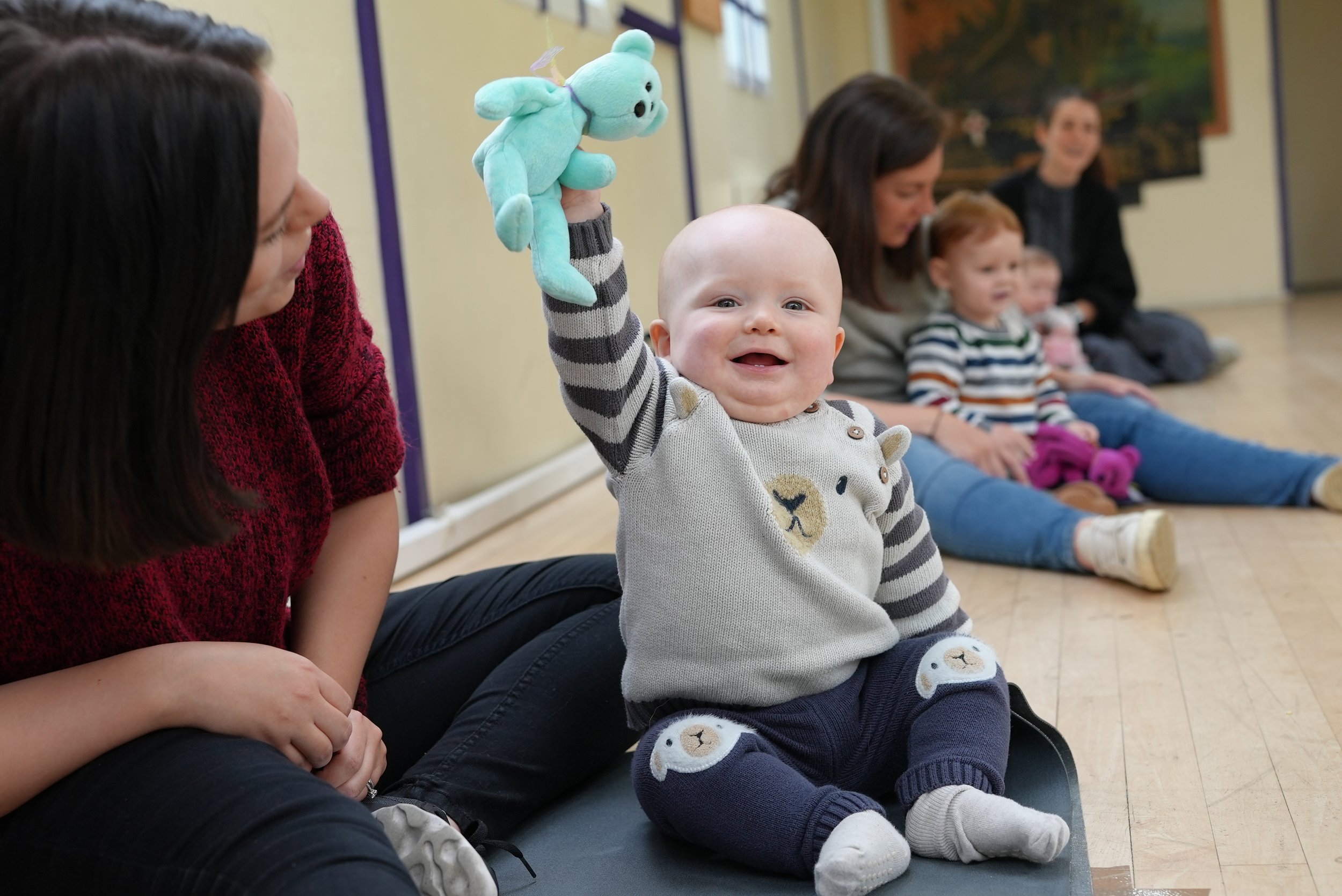  Little boy is smiling and holding up a blue teddy. Mum is sitting next to her baby smiling 