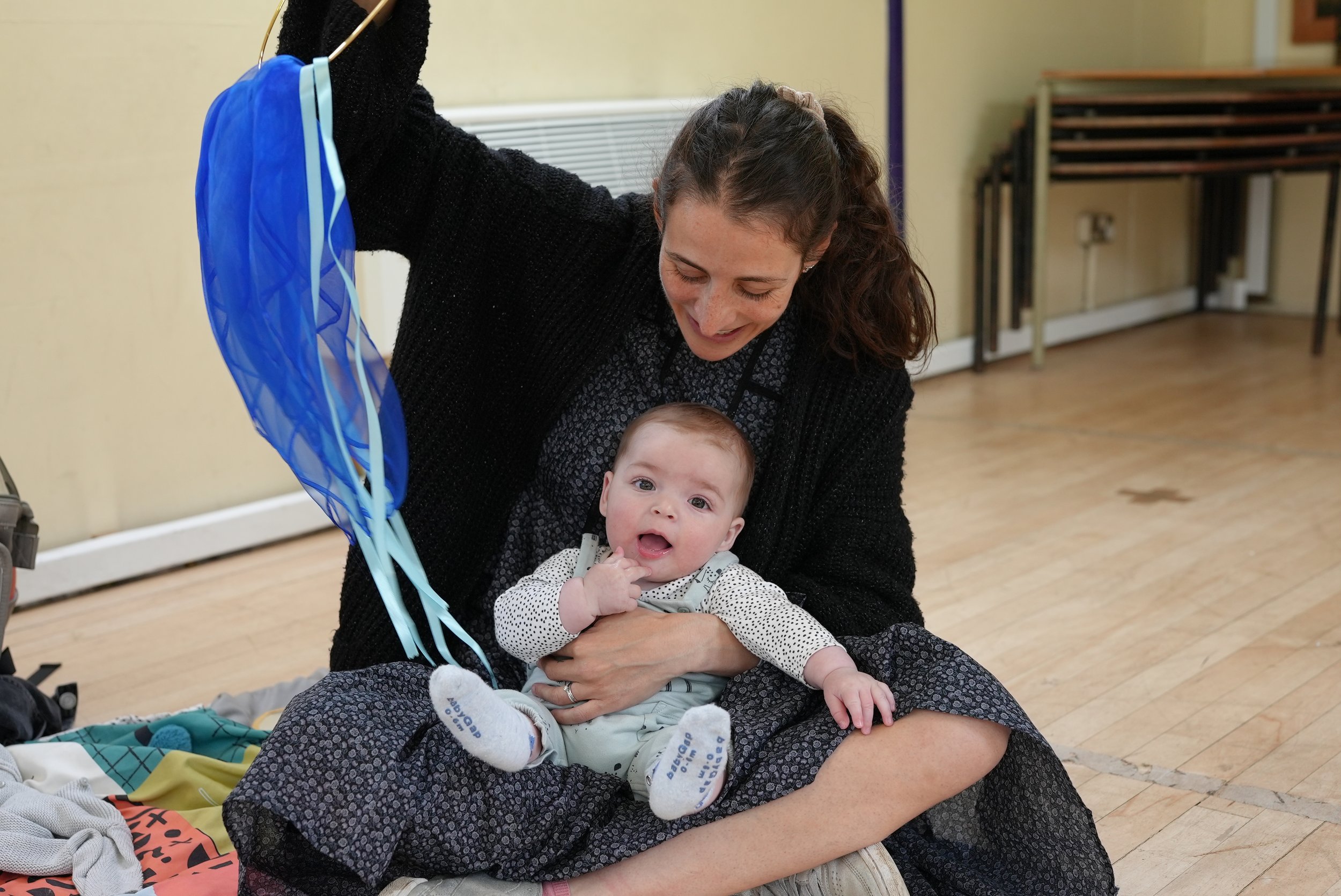  Little boy is sitting on mums lap. She is holding a blue silk scarf above him 