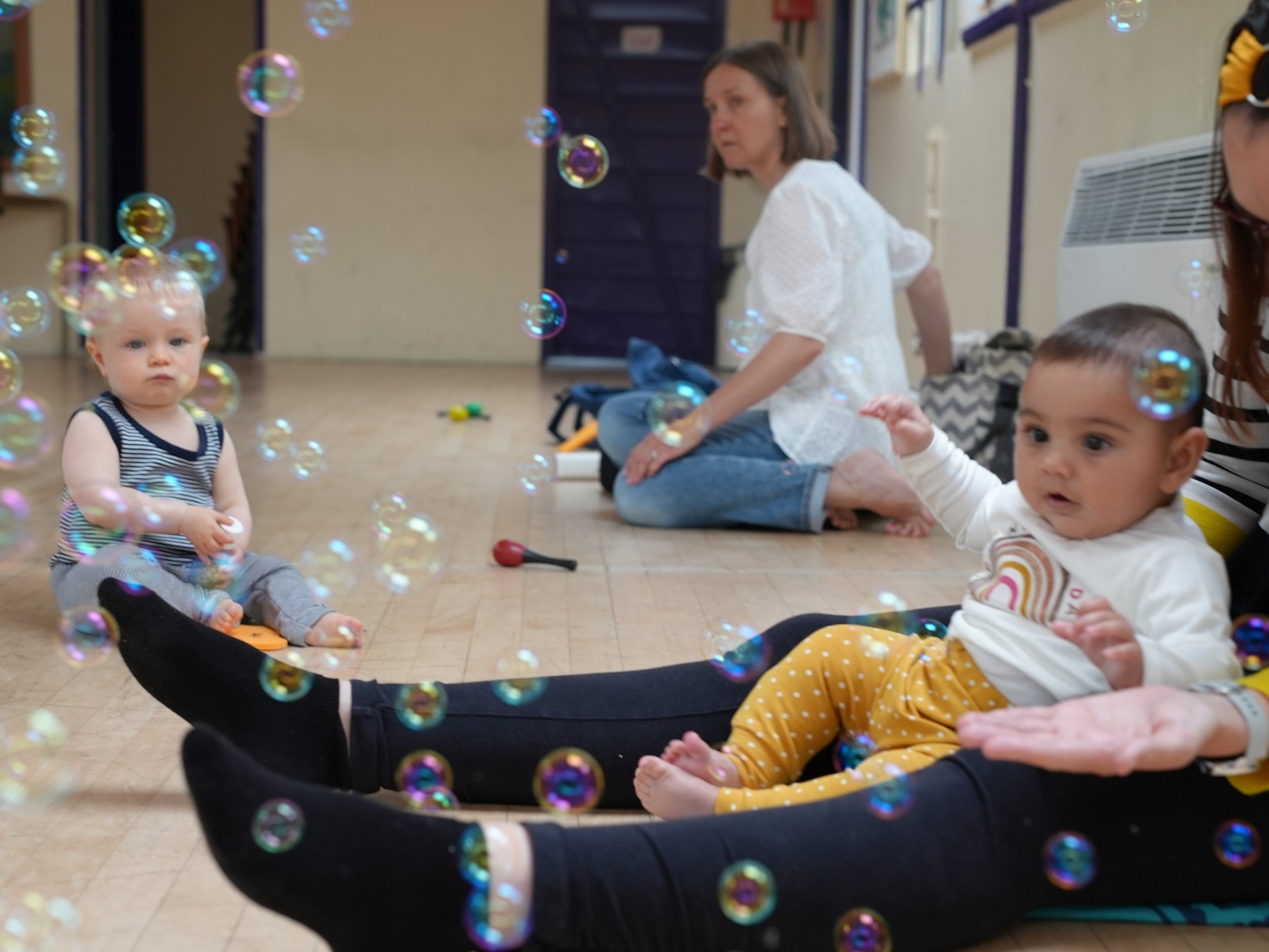  a little girl sitting in-between her mums legs is surrounded by bubbles  