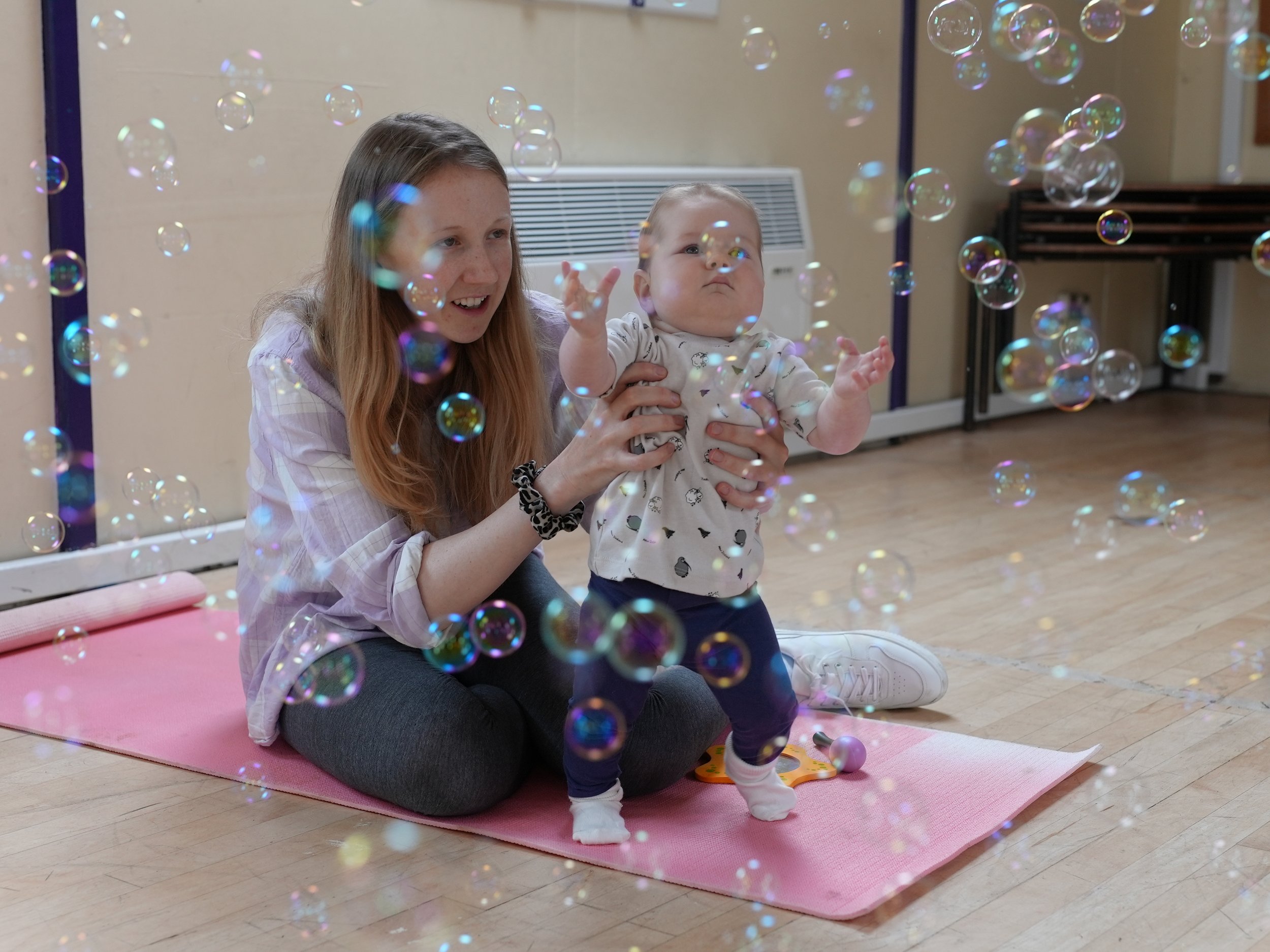  mum and baby girl are surrounded by lots of bubbles. The little girl is reaching out to pop them 