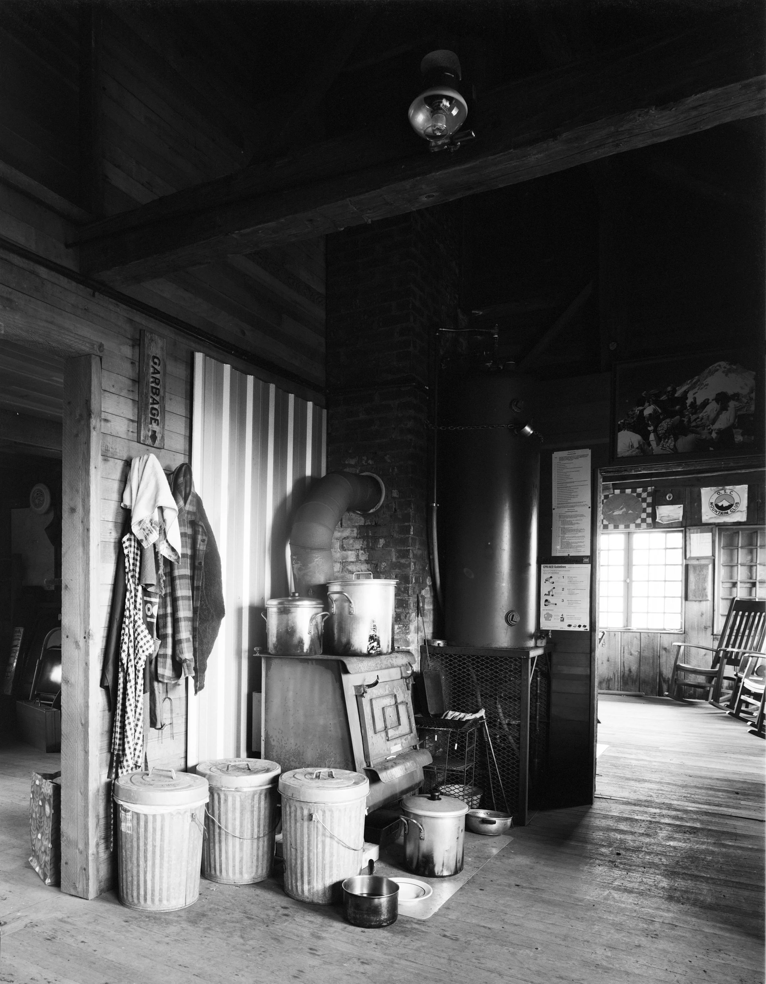 The wood burning stove is one of two in the building, the other sits in the dining room beyond on the opposite side of this chimney. Covered pails hold combustible waste, compost, and recyclables.