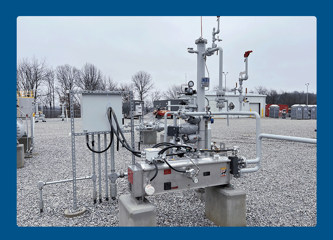 Industrial equipment with pipes, gauges, and control panels set outdoors on a gravel surface, surrounded by portable toilets and leafless trees.