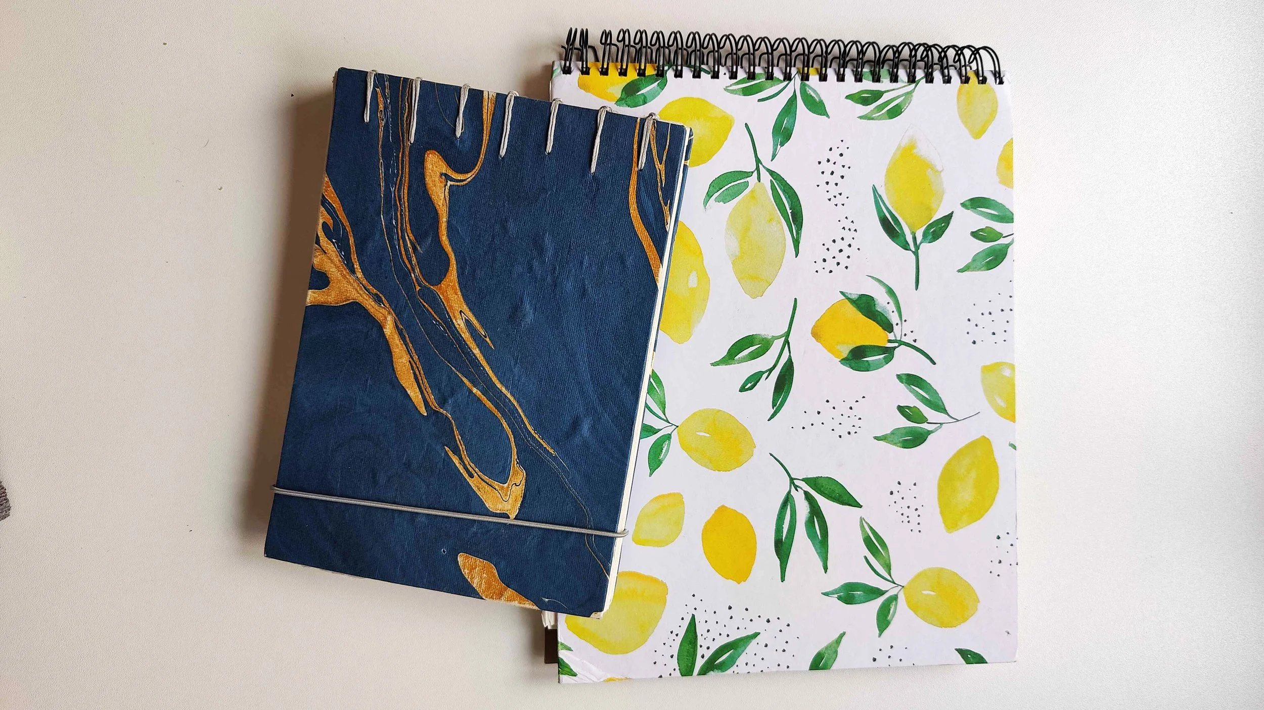 Two art journals stacked together, one with a blue and gold marbled cover and one with a lemon pattern cover, used for watercolour practice.
