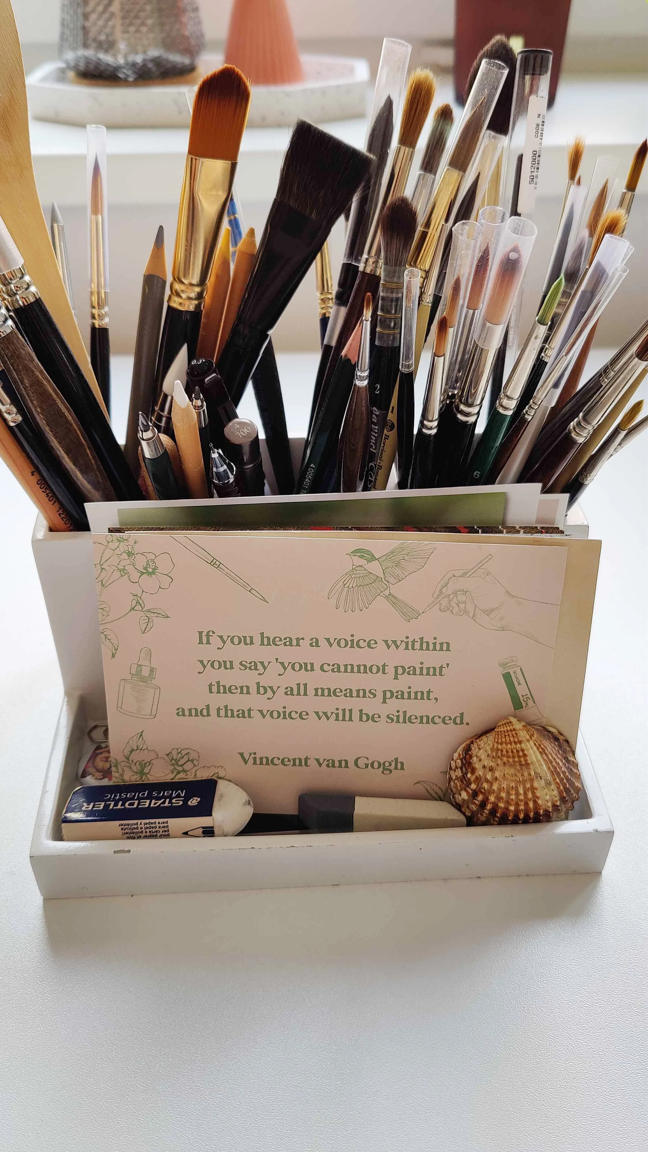 Container filled with watercolour brushes, pencils and tools on a desk with an inspirational Vincent van Gogh quote card.