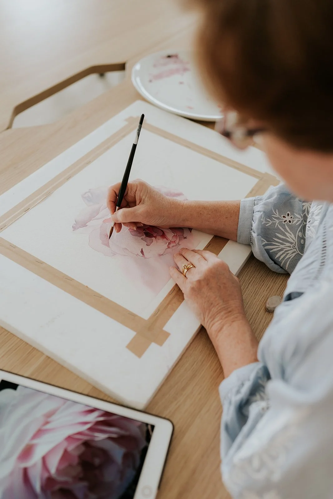 Artist (Louise De Masi) painting a soft pink flower in watercolour on taped paper, using a reference photo on a tablet, demonstrating step by step technique in an online class.