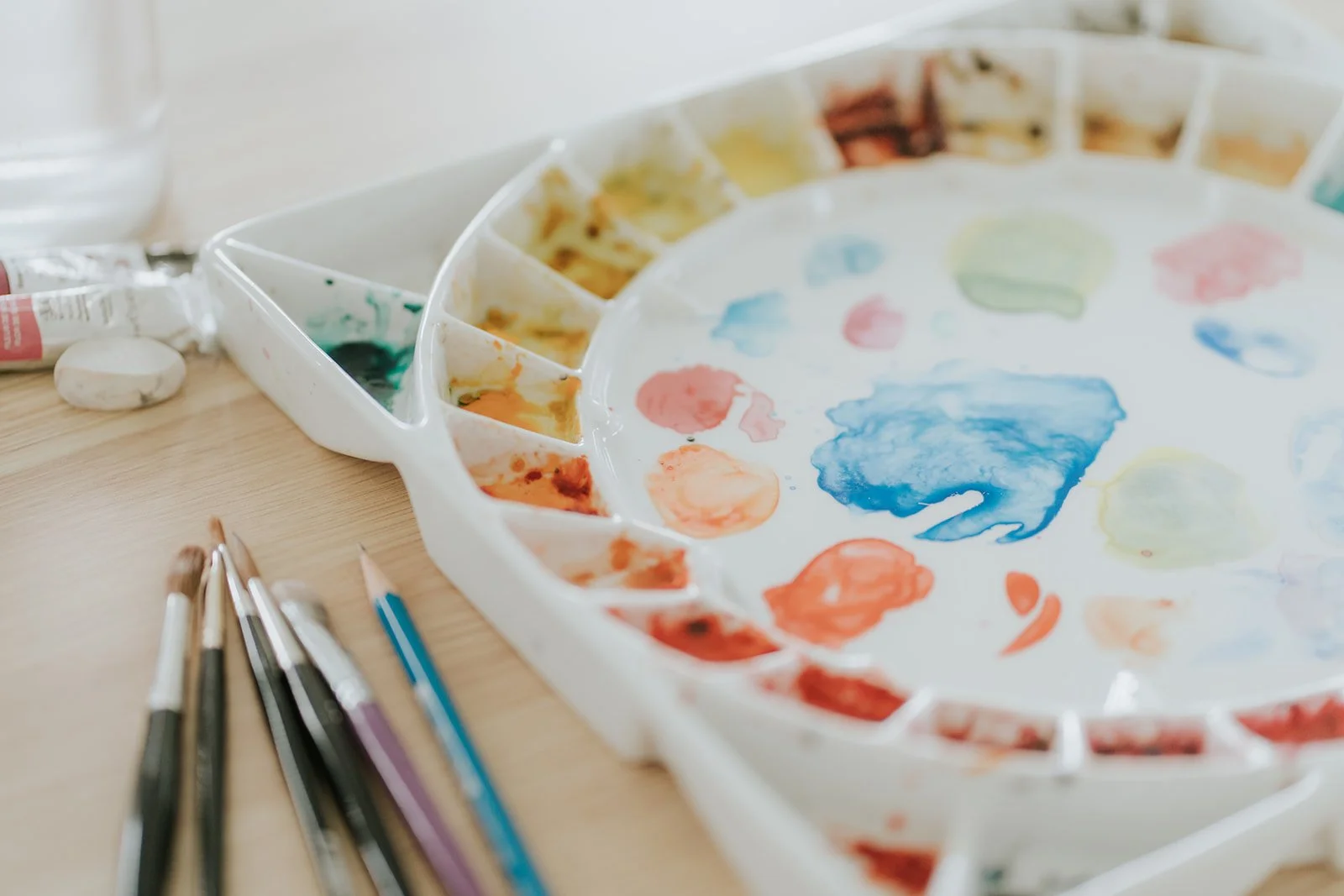 A well-used watercolour palette with mixed colours and several brushes and pencils resting beside it on a wooden table.