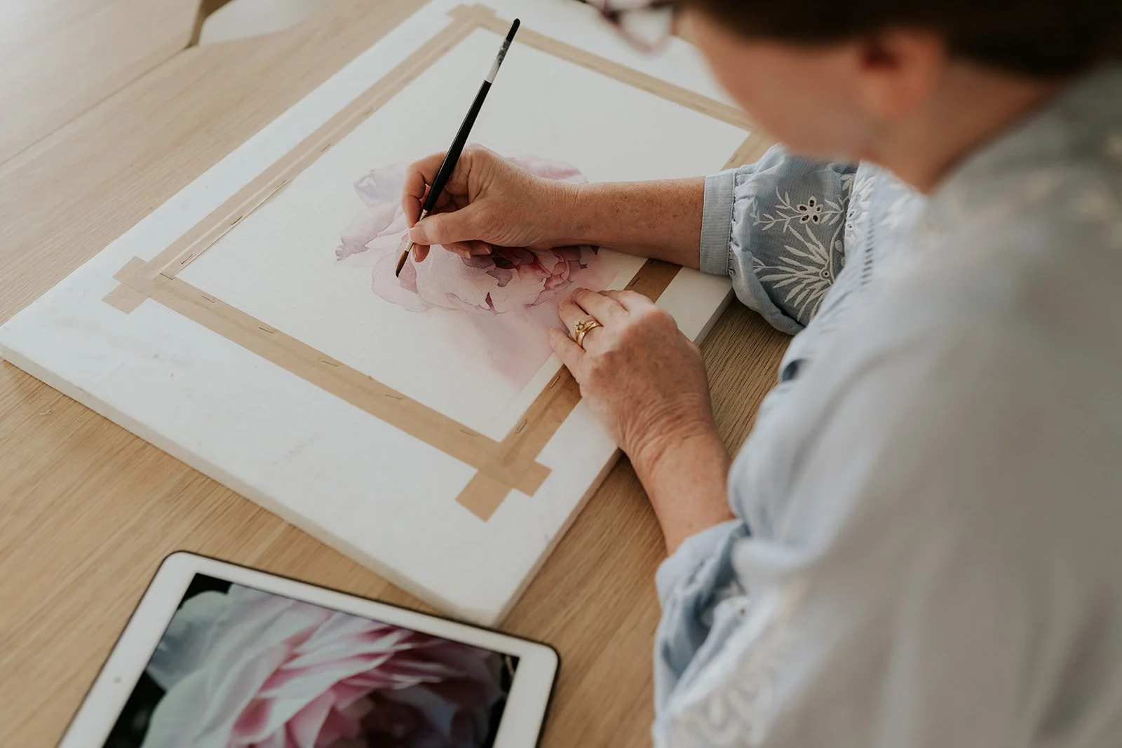 Louise De Masi carefully painting a soft pink rose in watercolour using a photo reference displayed on an iPad.