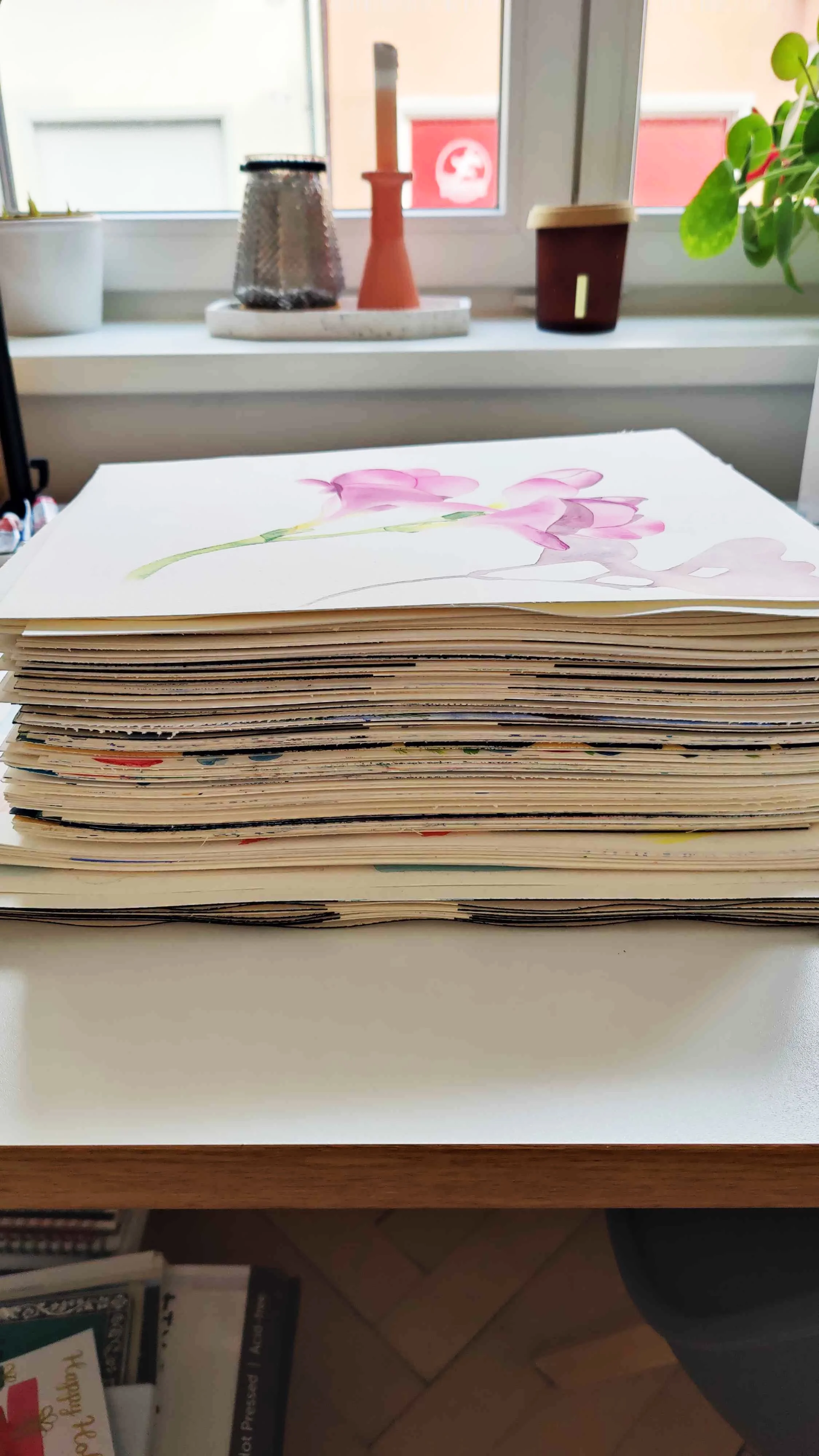 Large stack of watercolour practice sheets on a desk with a pink floral painting on top near a window.