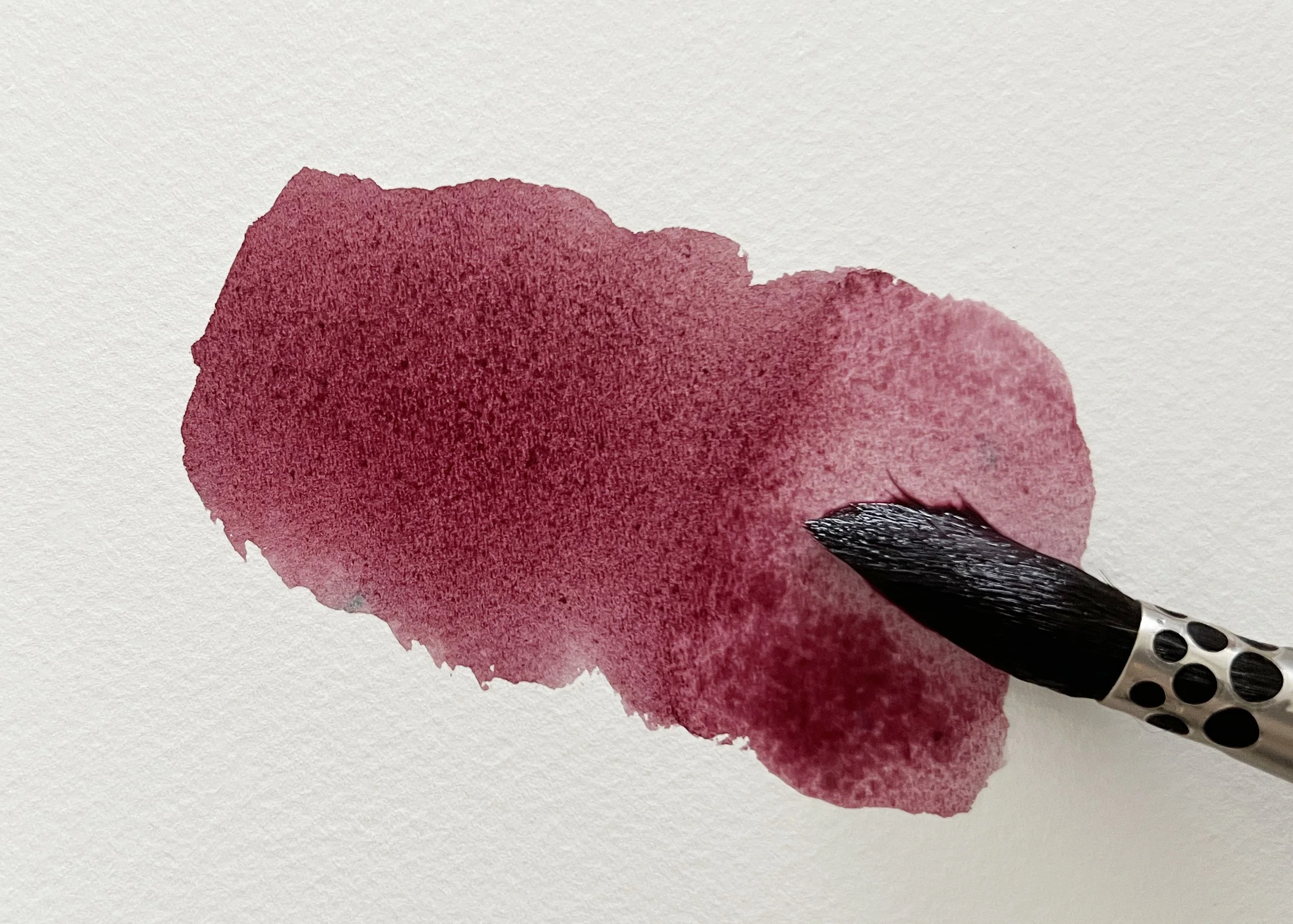 A rich plum red watercolour swatch in progress, with a brush blending out the pigment on cold-pressed paper.