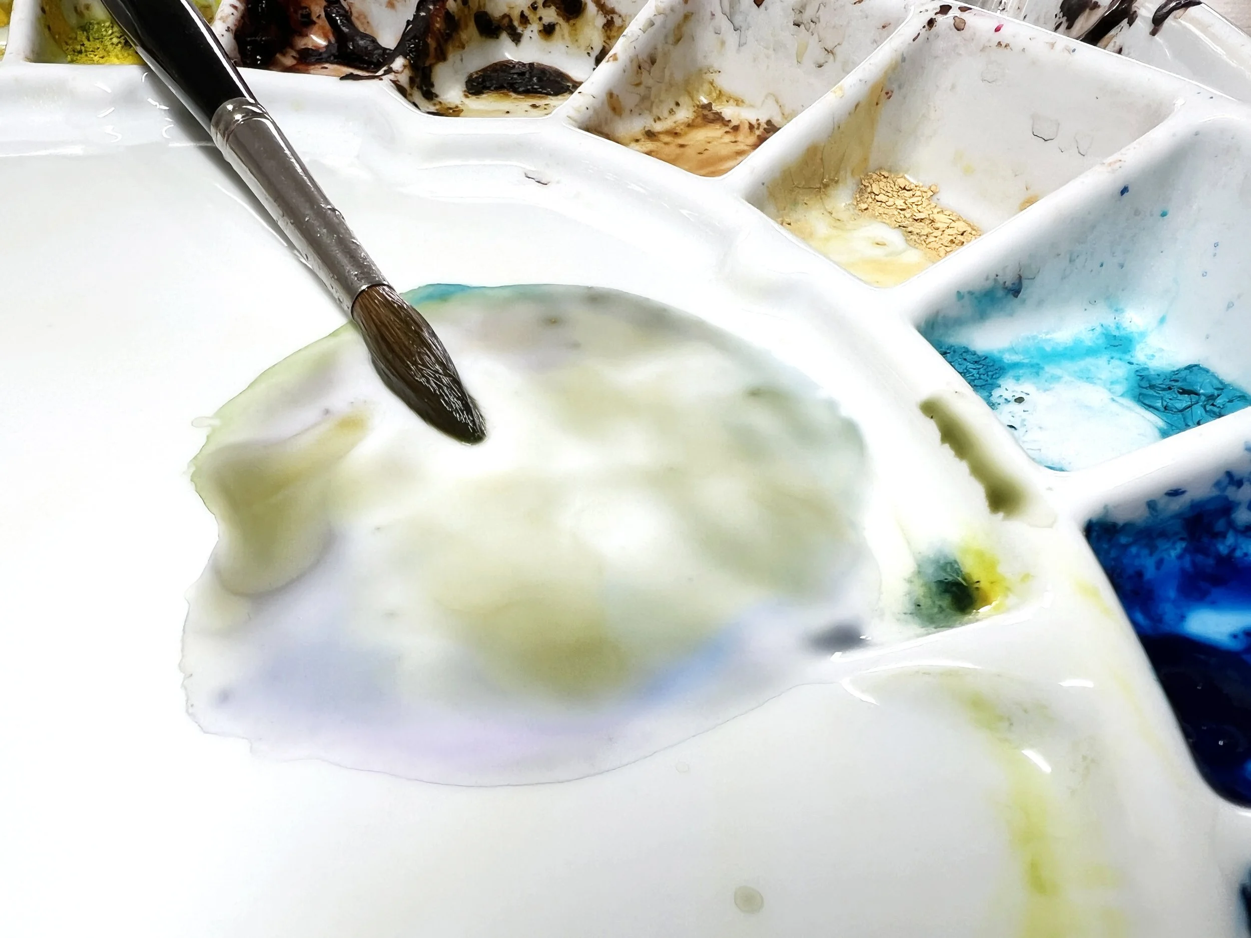 Round brush mixing a muted green watercolour wash on a ceramic palette with surrounding colour wells.