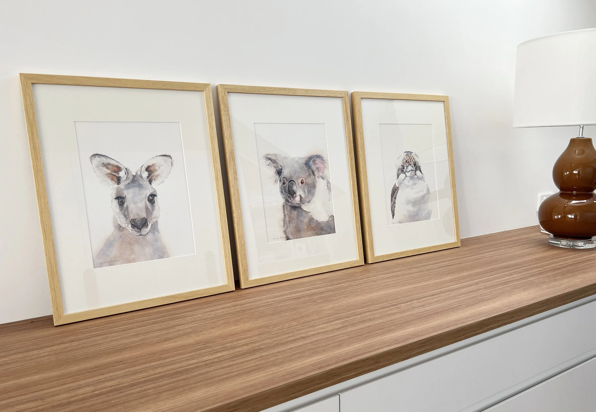 Three framed watercolour portraits of Australian animals – a kangaroo, a koala, and a kookaburra – displayed on a wooden cabinet under soft lighting.