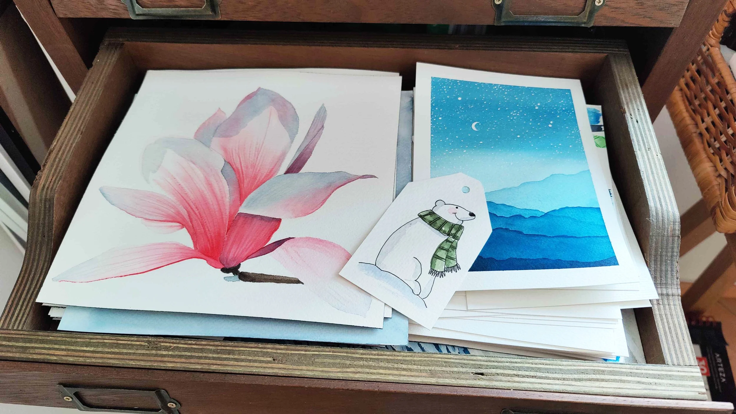 Open wooden drawer filled with finished watercolour paintings including a pink flower, night sky landscape and small polar bear illustration.