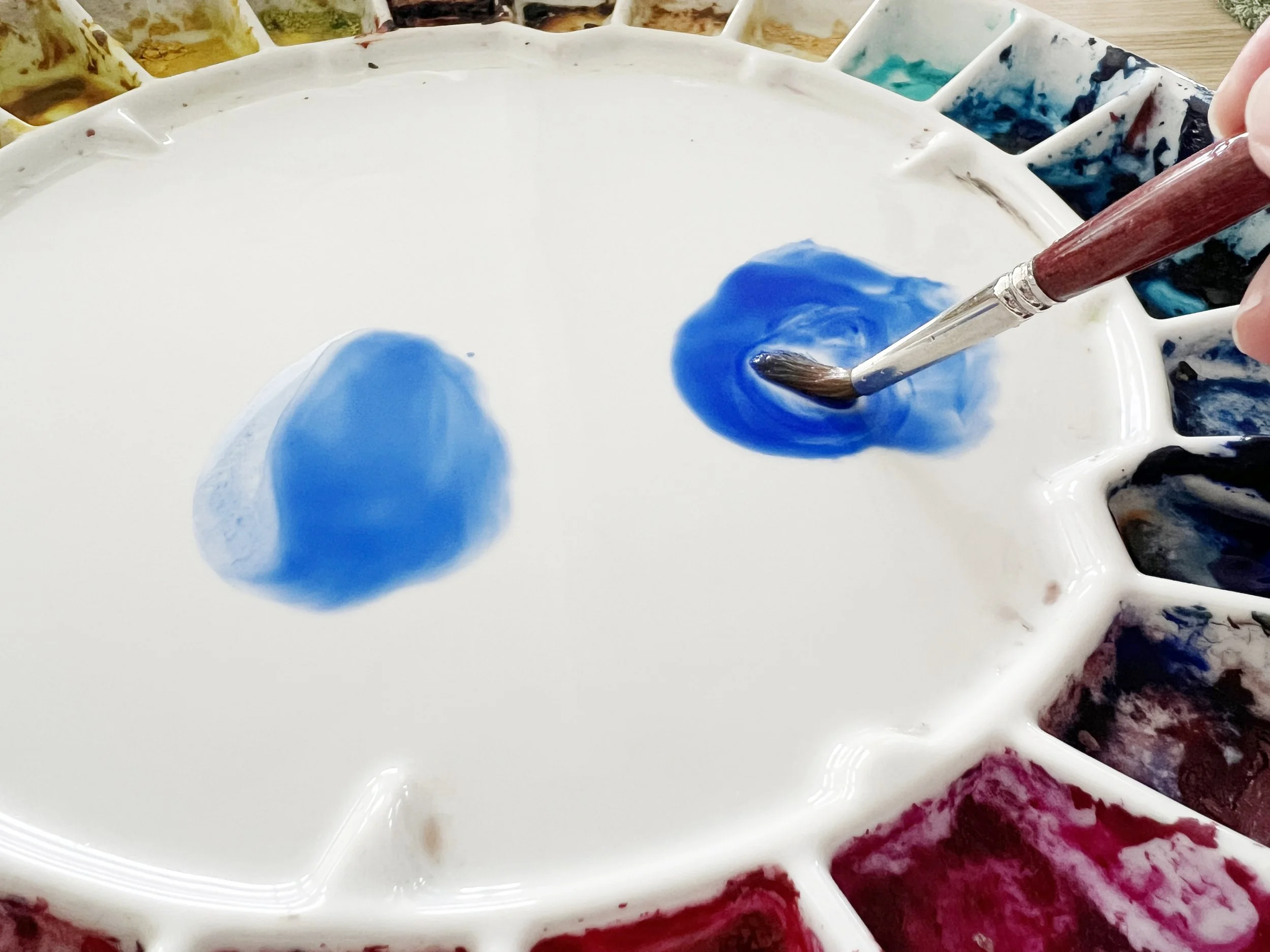 Close-up of a watercolour brush mixing concentrated blue pigment on a ceramic palette beside a lighter, more diluted blue mixture.