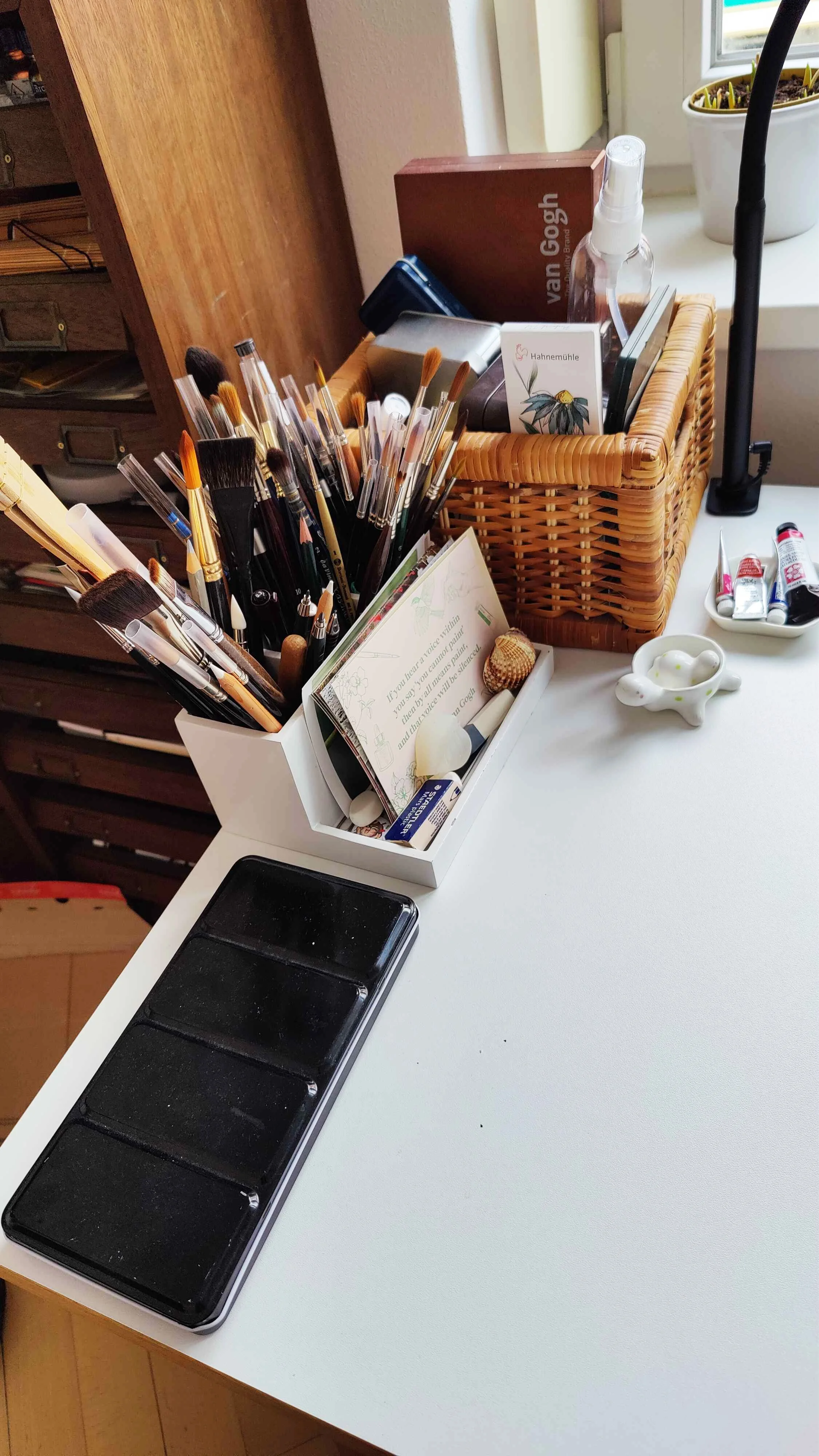Watercolour workspace with brush holder, wicker storage basket, paint tubes and palette arranged neatly on a white desk near a window.