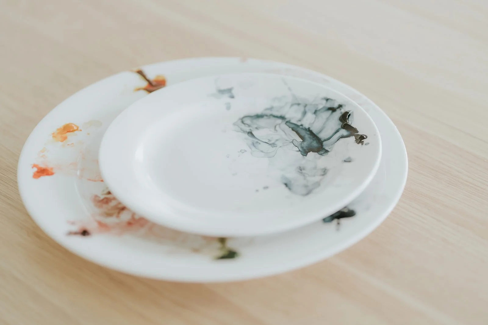 Two white ceramic plates stacked together, stained with dried watercolour paint in soft blacks, browns and reds.